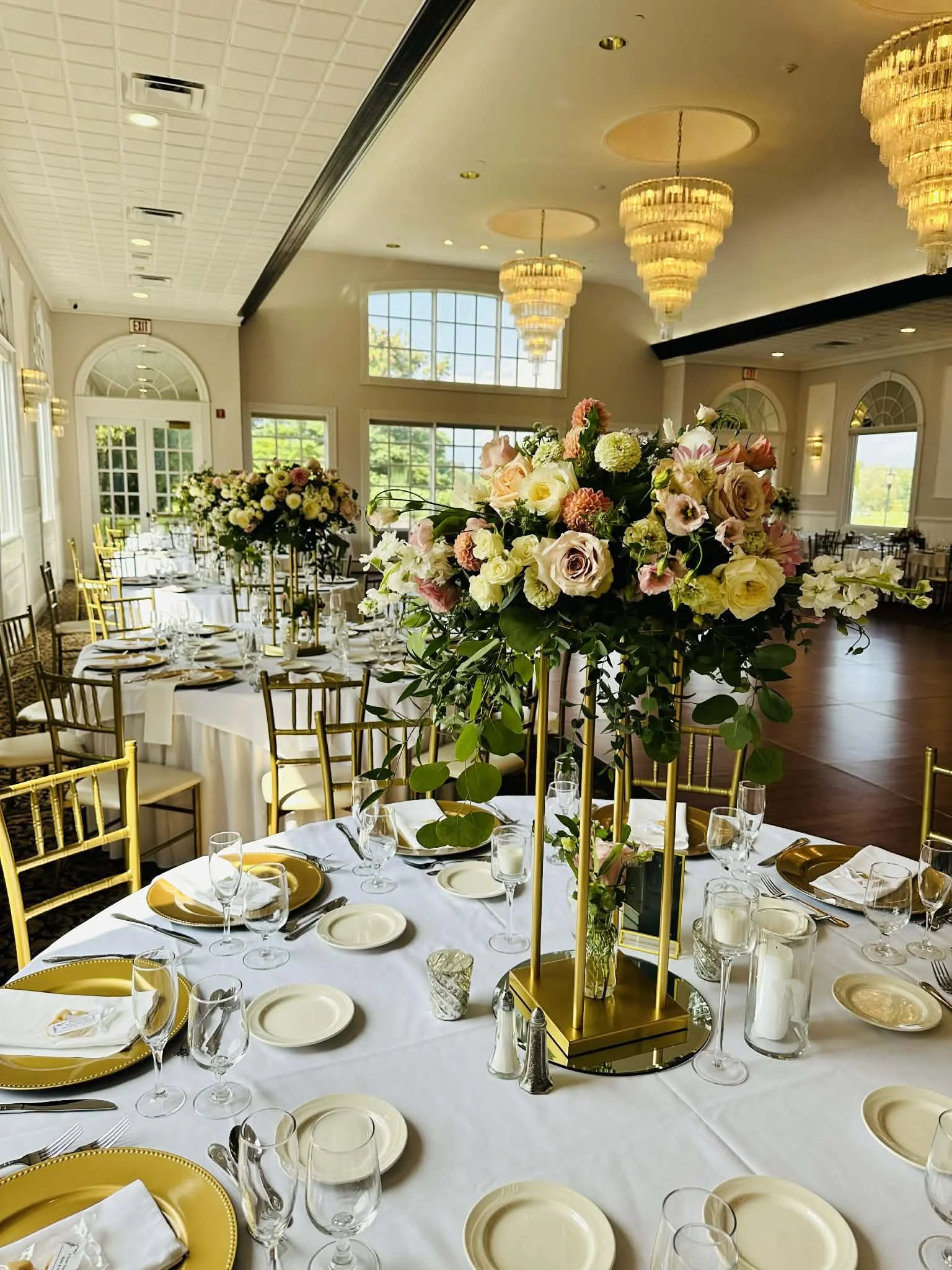 Elegant banquet table setup with a tall floral centerpiece in a bright, spacious room with large windows and chandeliers.