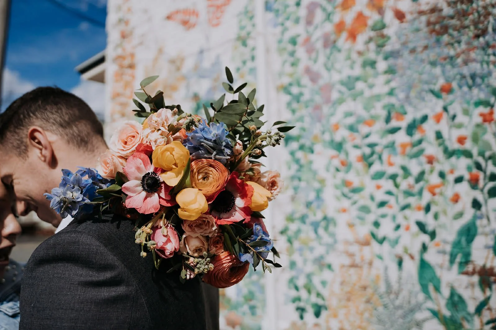 A man in a dark suit holding a large colorful bouquet of flowers, standing outdoors in front of a vibrant, completion or floral mural.