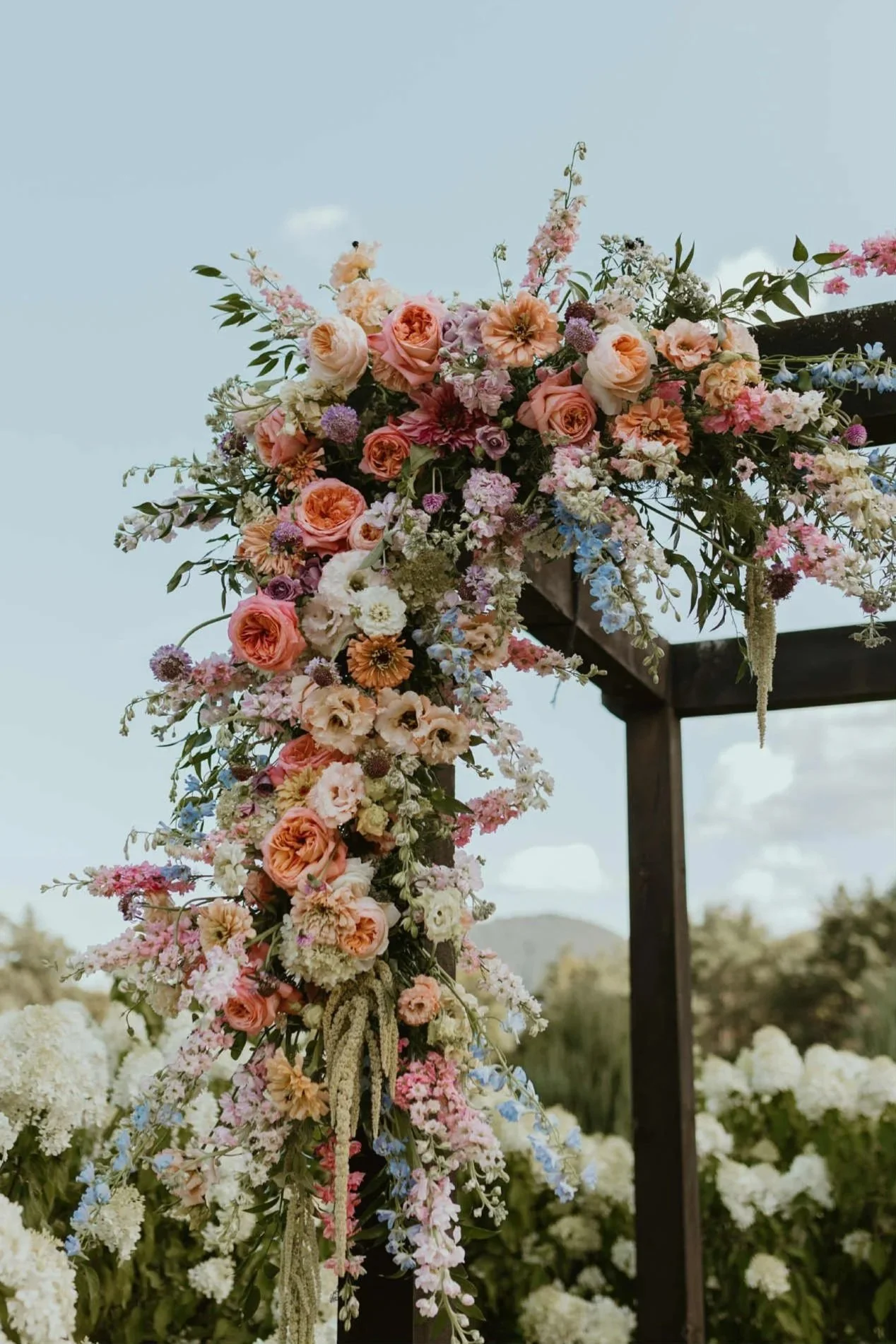 A floral wedding arch decorated with pink, peach, purple, and white flowers, set outdoors against a cloudy sky.