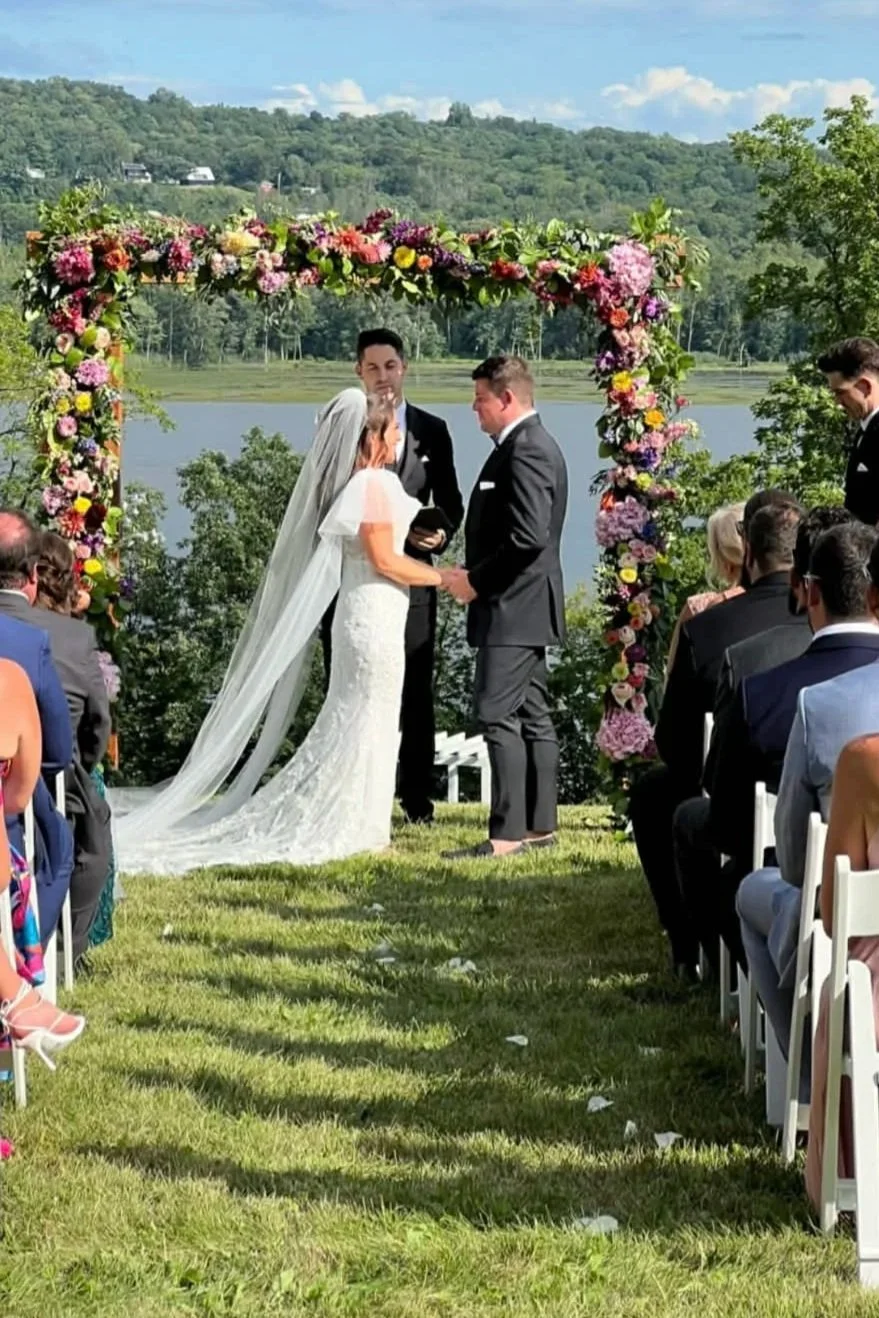 A couple getting married outdoors under a floral arch made by Lot 32 Flower Farm with a lake in the background and guests seated on either side.