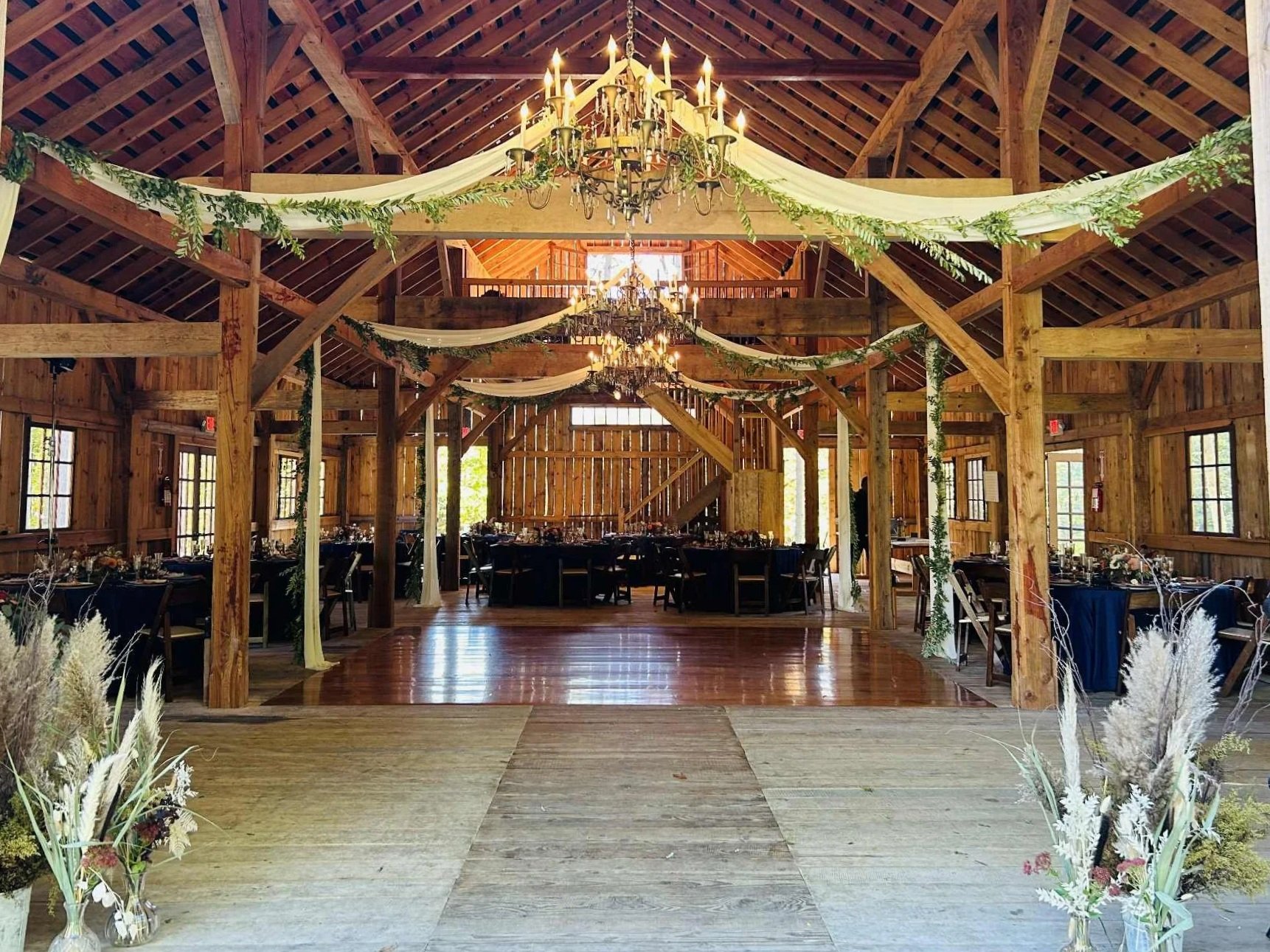 A rustic wedding reception hall decorated with greenery and white drapes, featuring wooden beams, chandeliers, and round tables with chairs.