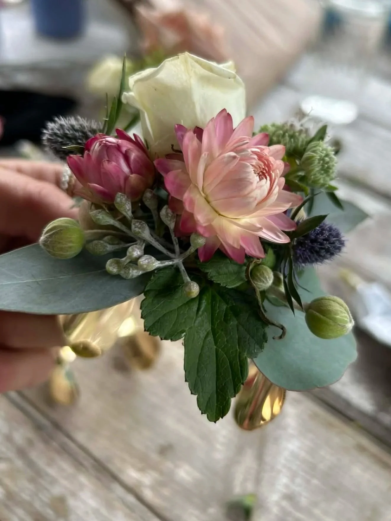 A small floral arrangement featuring a white rose, pink dahlias, green foliage, and purple and green buds, held in a copper-colored container.