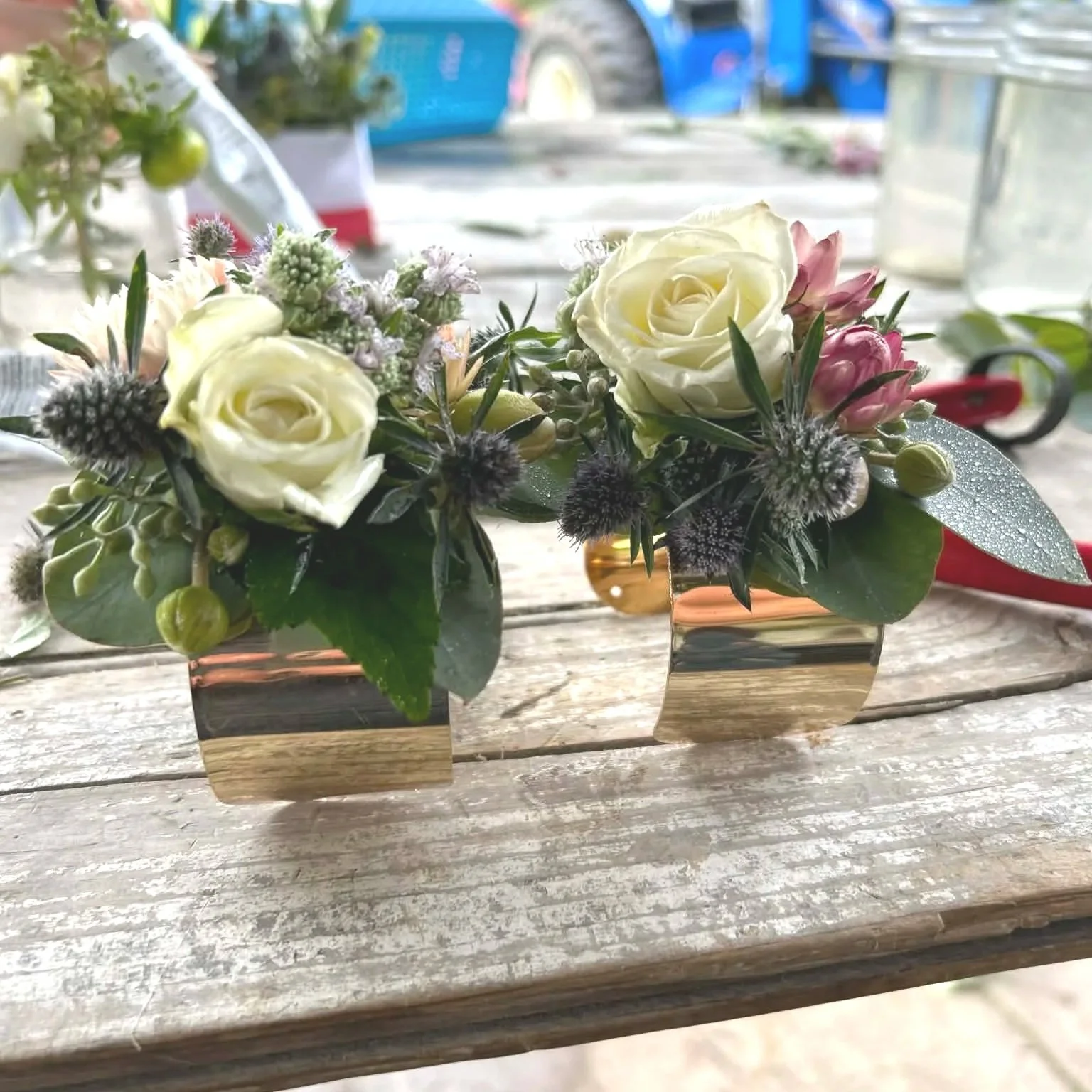 Two small floral arrangements with white roses, pink flowers, and various greenery in small gold and copper-colored containers on a wooden surface.