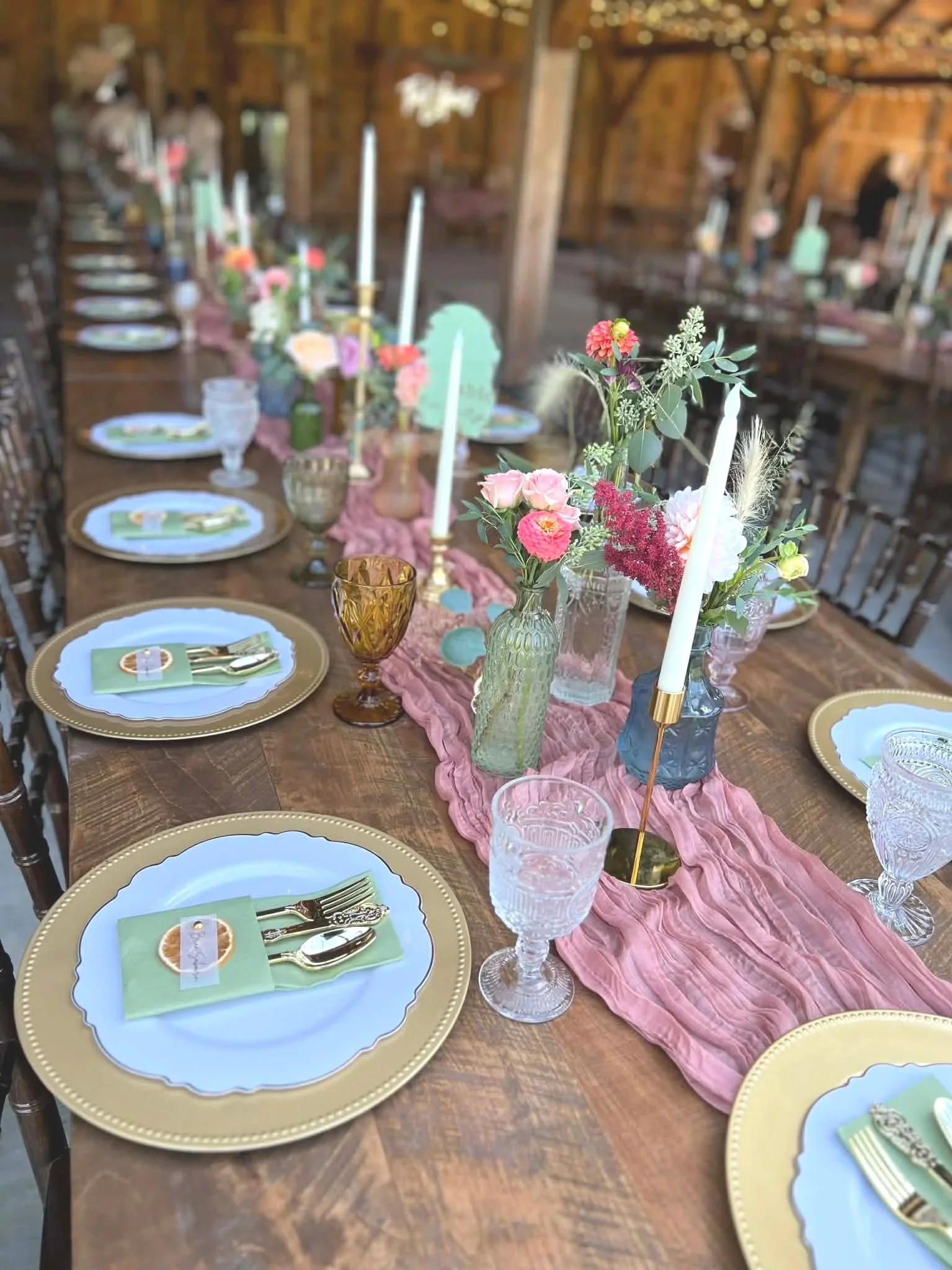 Table set for a celebration with plates, cutlery, and colorful vases filled with pink and red flowers, a pink table runner, and tall white candles in gold holders inside a rustic barn.