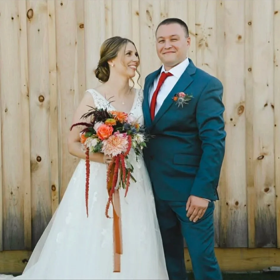 A bride and groom standing together in front of a wooden fence, the bride holding a colorful bouquet, both smiling and dressed in wedding attire.