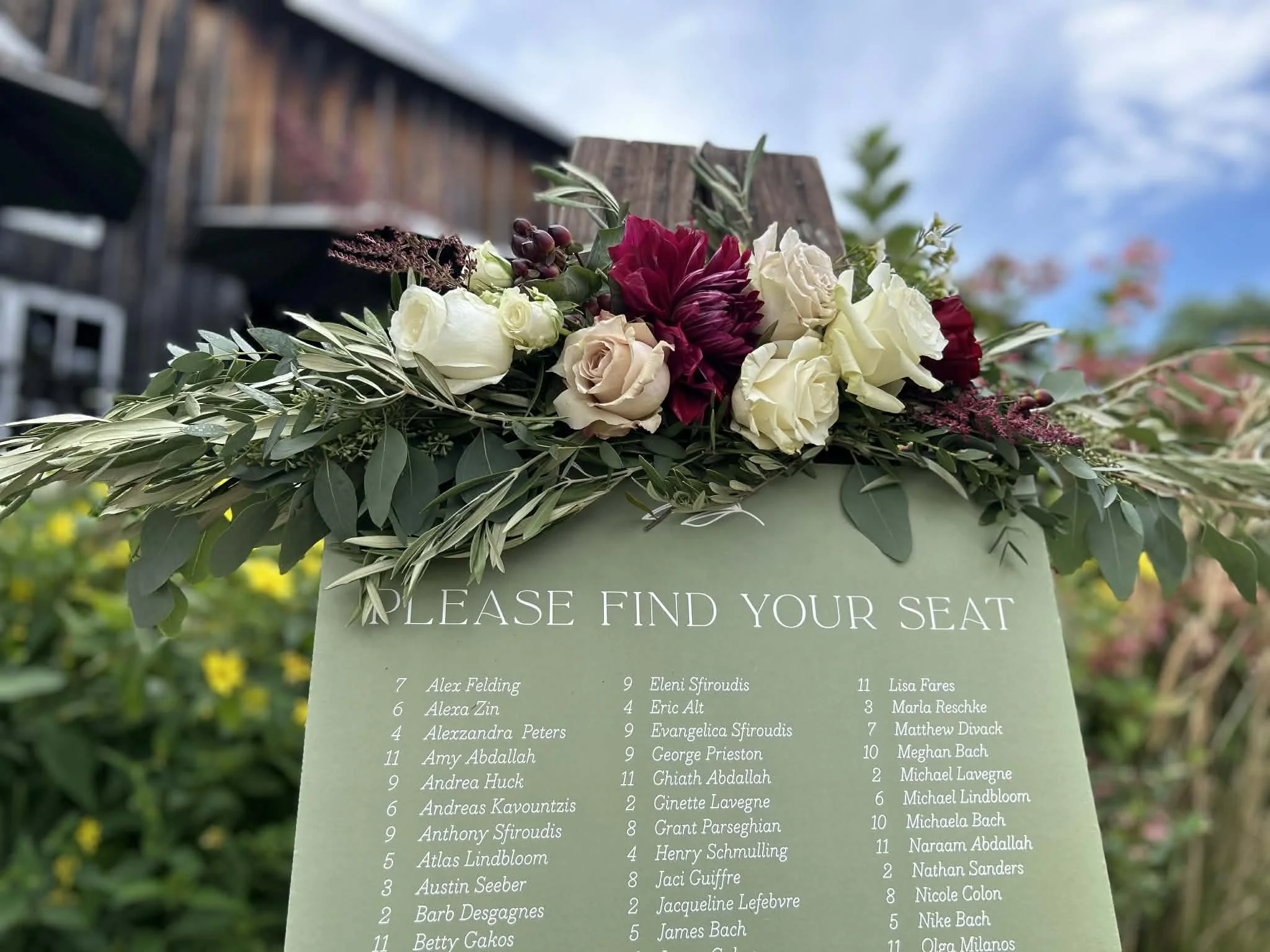 Wedding seating chart with a floral arrangement on top, featuring roses, and greenery. The chart lists guest names with table numbers, with the sky and some flowering trees in the background.