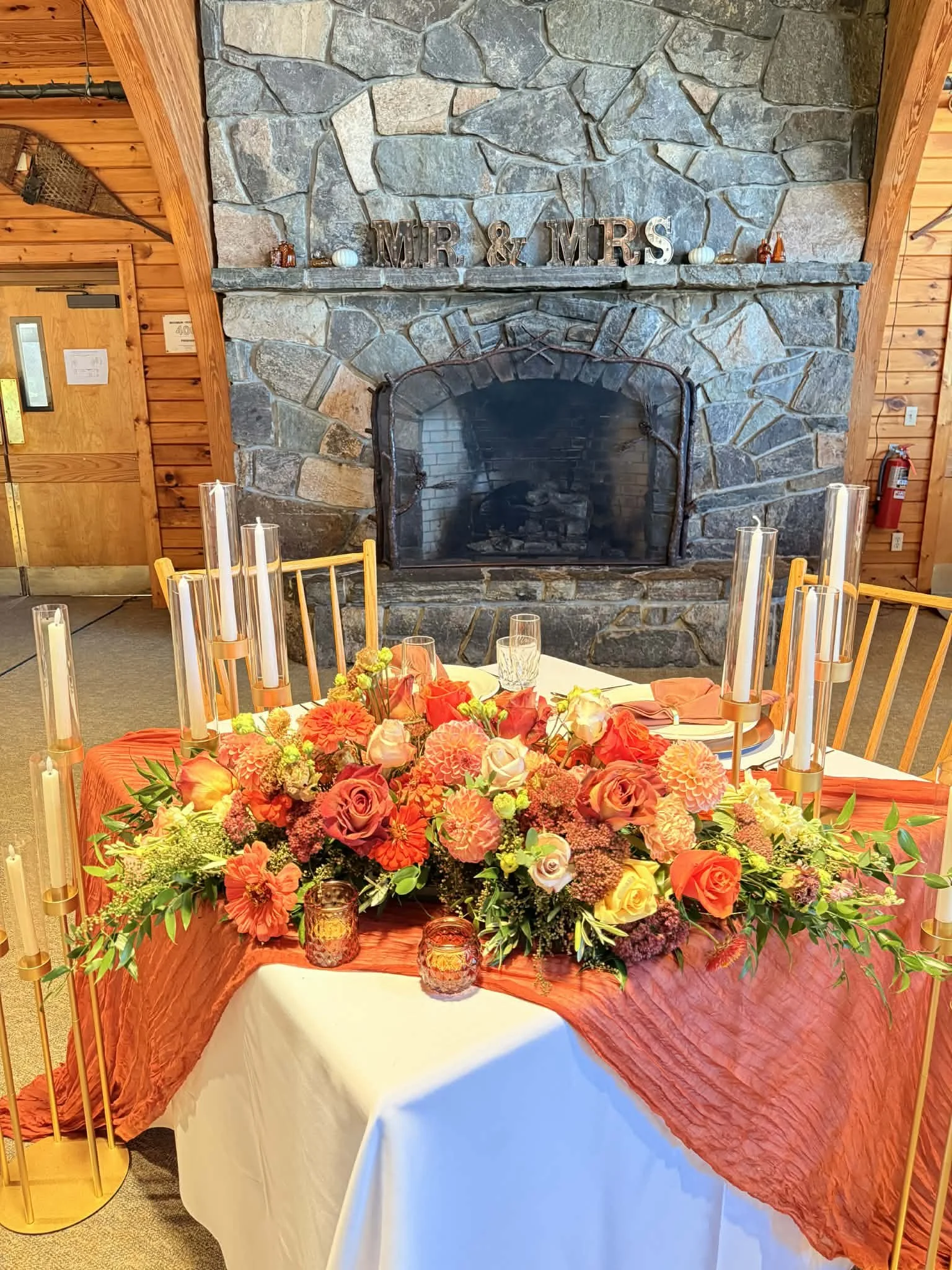A decorated dining table with a floral centerpiece and candles in front of a stone fireplace with a mantel that says 'MR & MRS' in a warmly decorated room.