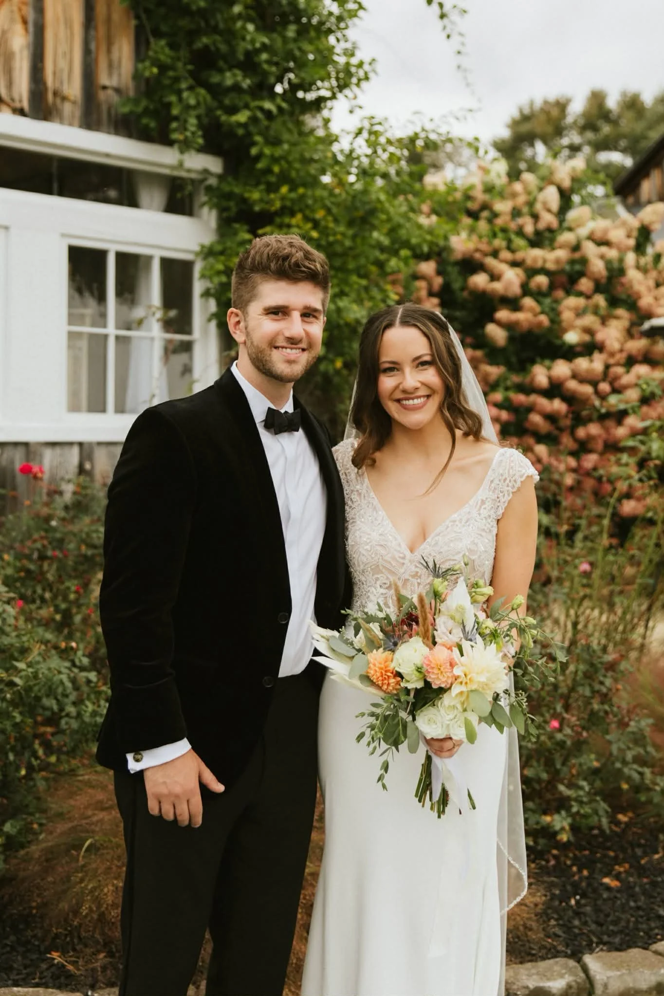 A bride and groom standing outdoors in a garden, smiling for a wedding photo. The bride is holding a bouquet of flowers and wearing a lace wedding dress with a veil. The groom is dressed in a black tuxedo with a bow tie, and they are surrounded by gr