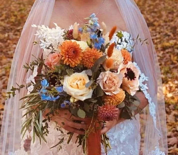 A bride holding a colorful bouquet of flowers, including white, peach, orange, blue, and pink blooms, with greenery, in an outdoor setting with fallen leaves on the ground.
