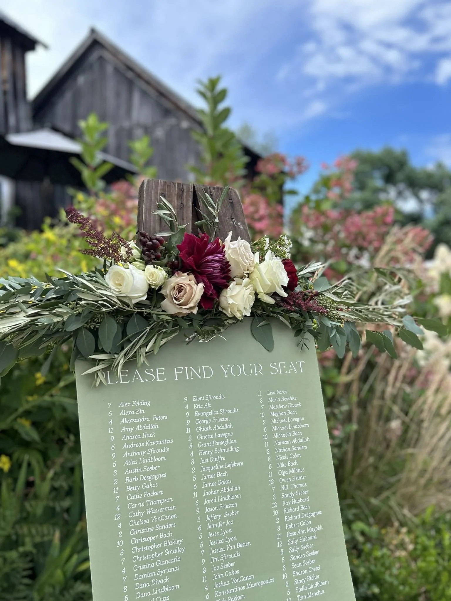 A wedding seating chart with a beautiful floral arrangement on top designed by Lot 32 Flower Farm, displaying names and table numbers. The background shows a rustic barn, trees, and a partly cloudy sky.