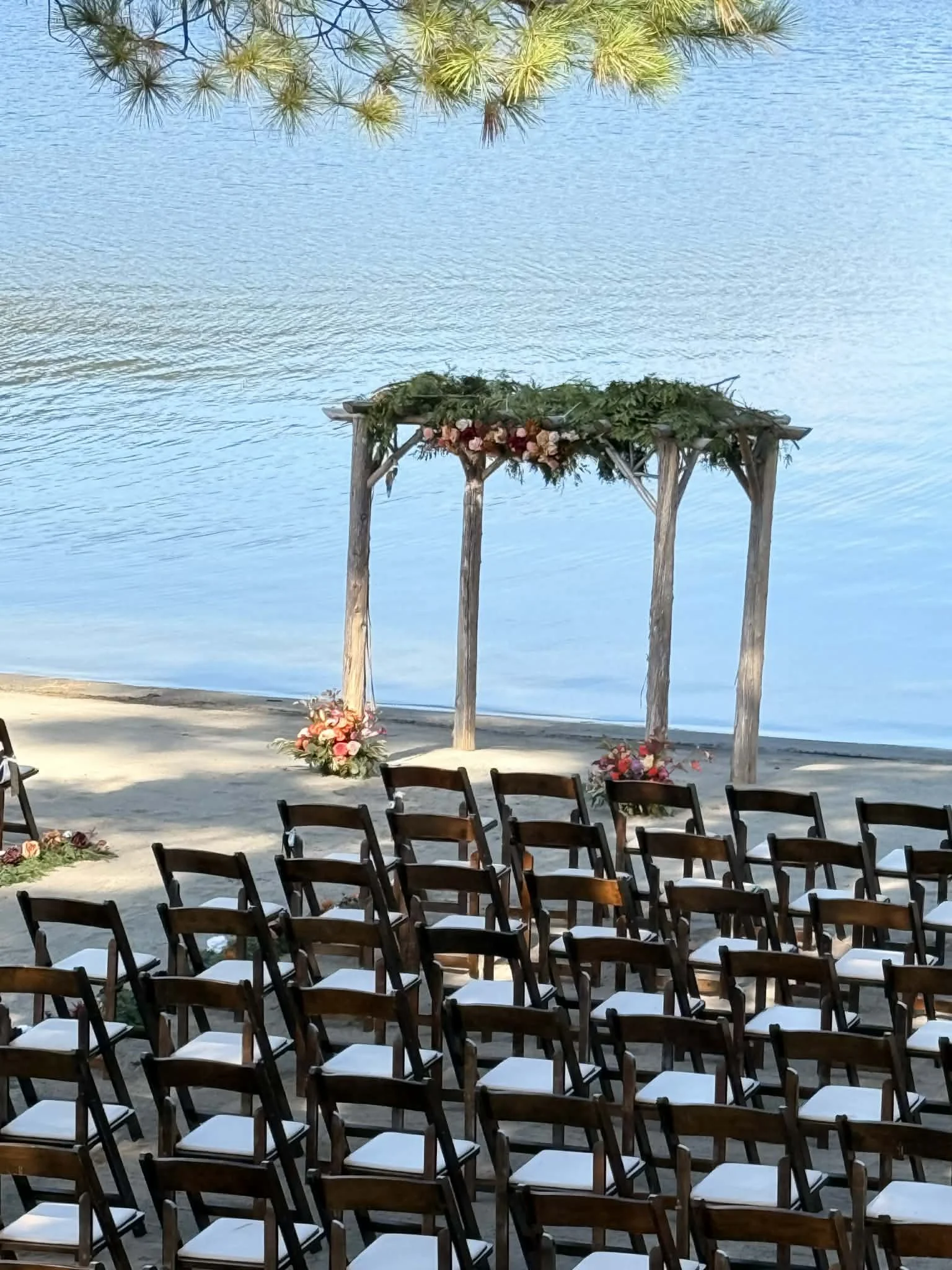 A beach wedding setup with wooden chairs facing an arch decorated with flowers and greenery, set on the sandy shore by a body of water.