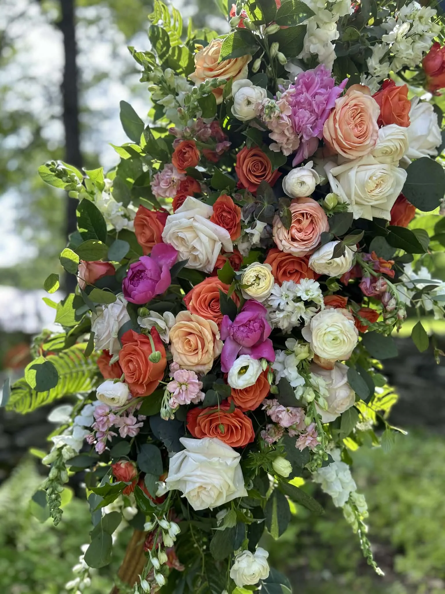 A large, colorful floral wedding arrangement featuring roses, peonies, and other flowers in shades of peach, white, pink, and orange, with green foliage background. This is part of a wedding arbor made by Lot 32 Flower Farm.