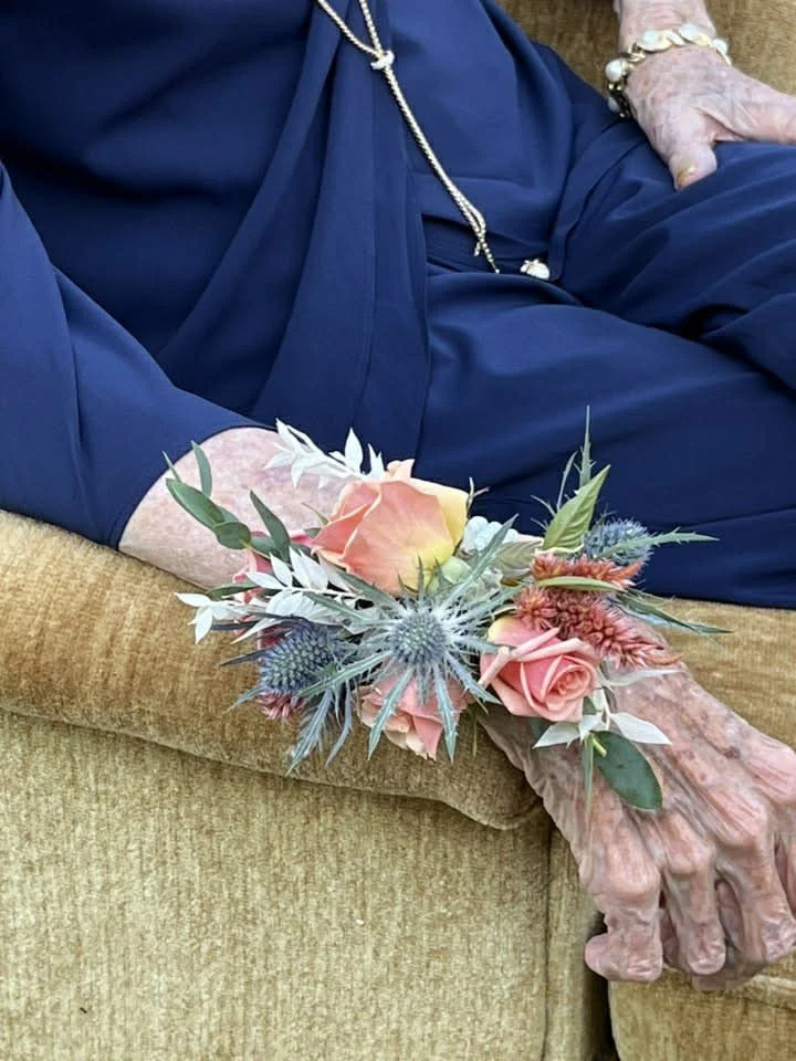 An elderly woman resting on a beige couch, holding a bouquet of peach and pink roses along with other flowers and greenery.