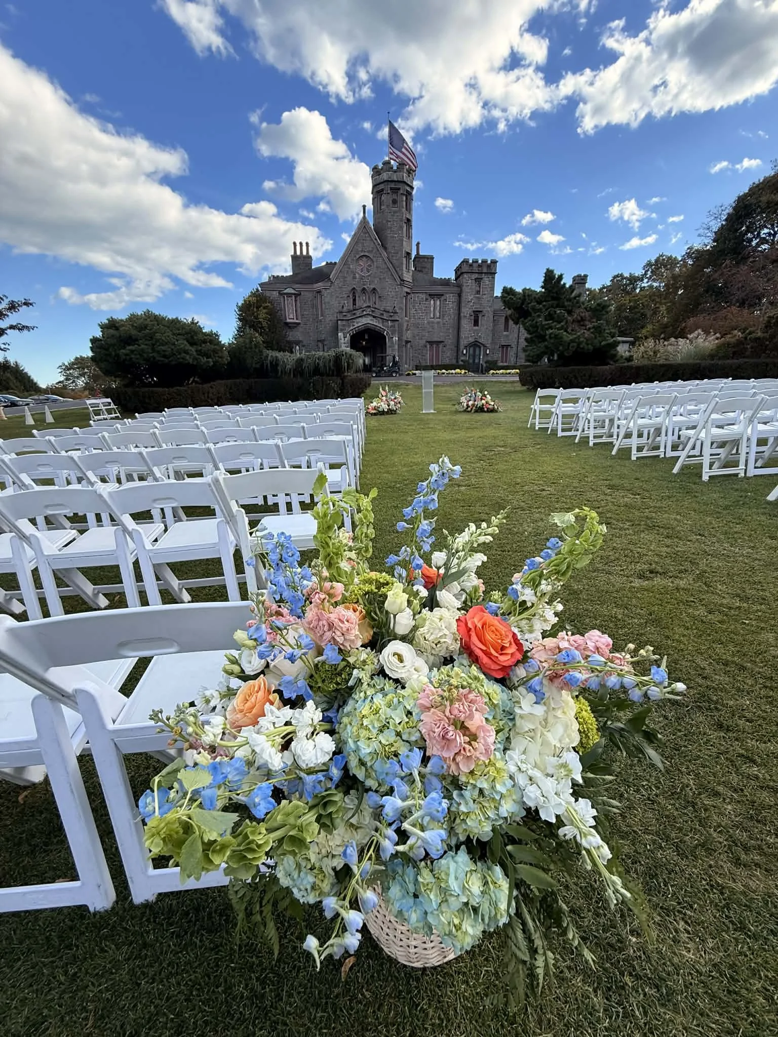 An outdoor wedding setup featuring white chairs arranged on either side of a grassy aisle, with a colorful flower arrangement in a basket at the front. In the background, a historic castle-like building with a tower and an American flag flying on top