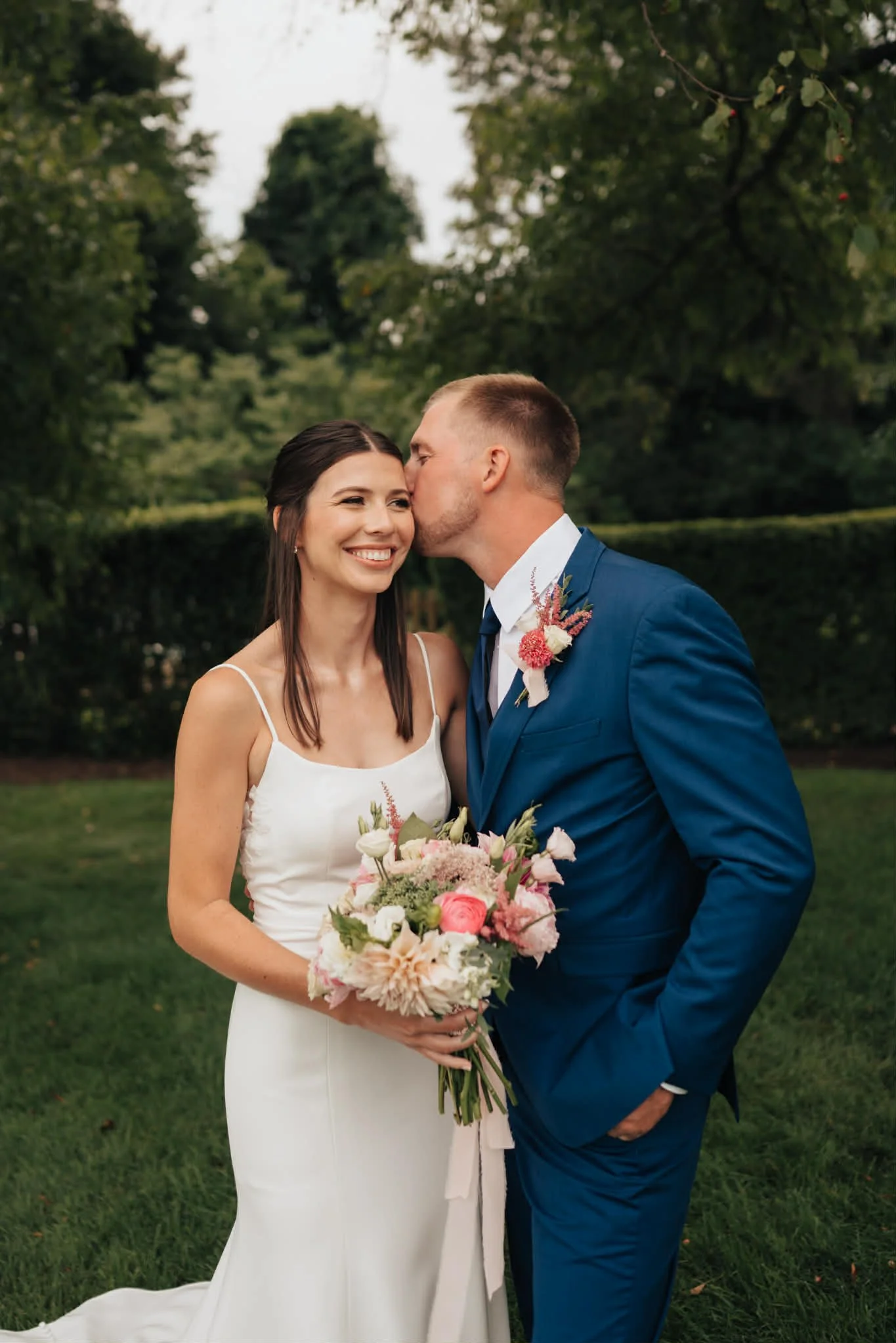 A newlywed couple outdoors with trees in the background. The woman in a white dress is smiling while the man in a blue suit kisses her on the cheek. She holds a bouquet of pink and white flowers.