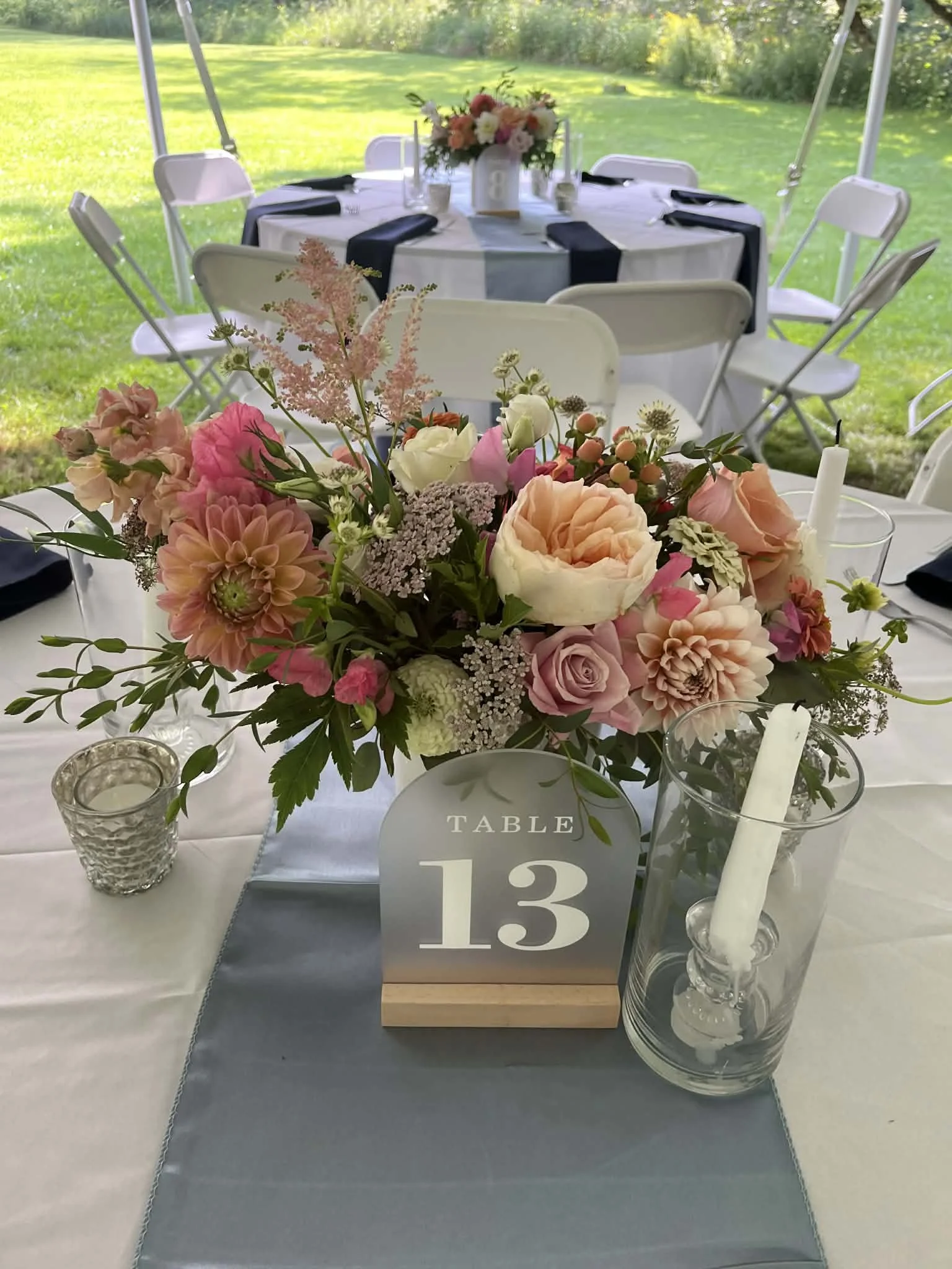 A table decorated with pink, white, and peach flowers, a pitcher with a candle, and a table number sign for table 13, set outdoors with greenery in the background.