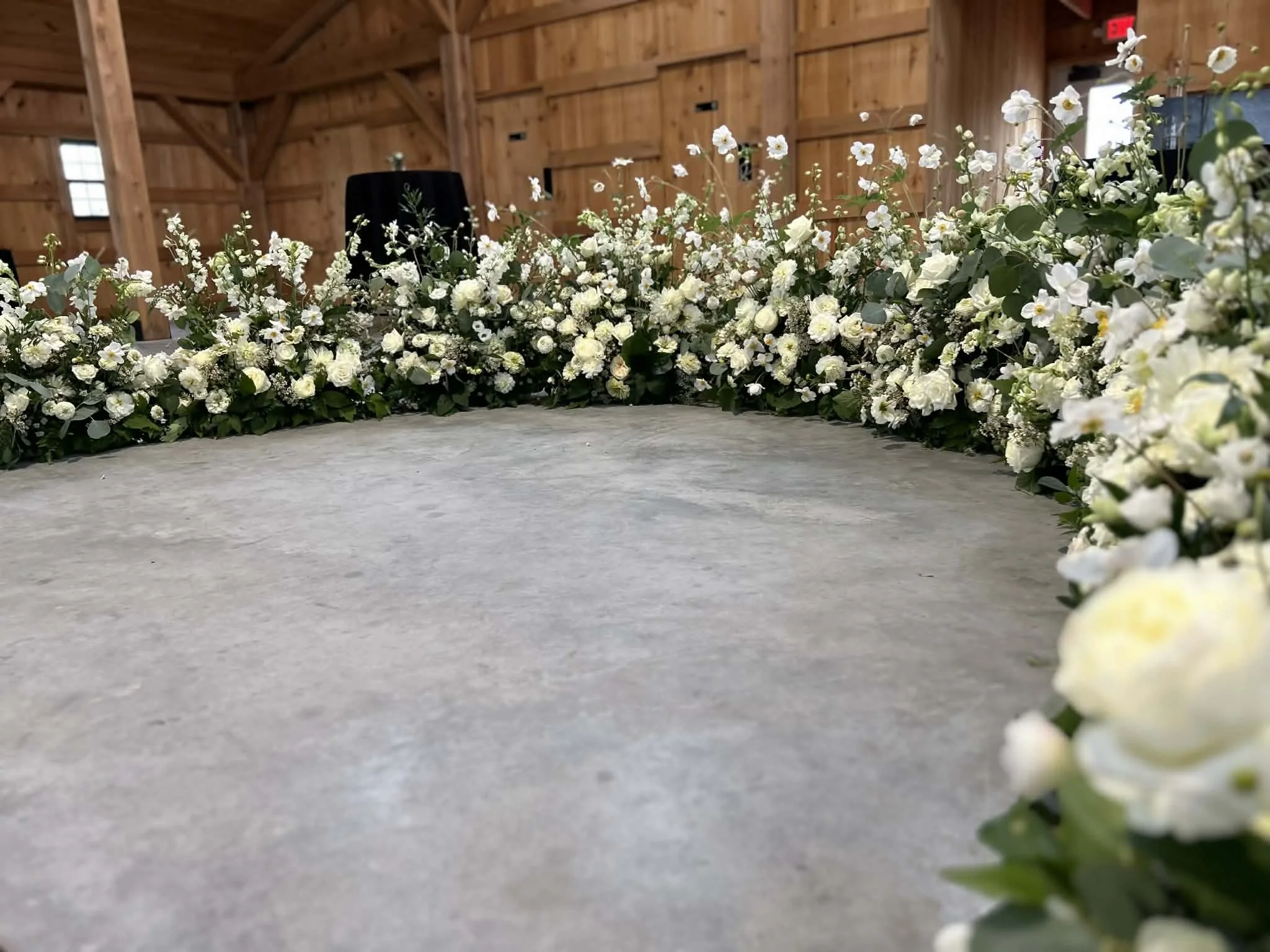 Floral arrangement by Lot 32 of white flowers and greenery in a semi circle around where the bride and groom will stand in a wooden barn setting.