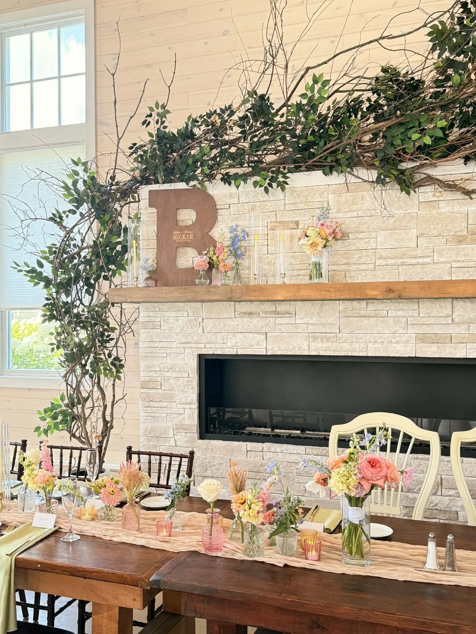 A decorated interior space with a long wooden table set with floral centerpieces, candles, and place settings, situated in front of a white brick fireplace with a wooden mantle. The mantle holds a large wooden letter 'B', flower vases, and candlestic