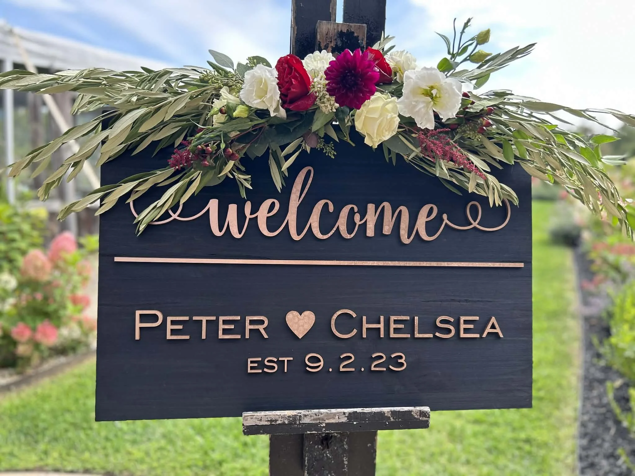 A dark-colored welcome sign with pink lettering, decorated with a floral arrangement by Lot 32 Flower Farm. of white, red, and purple flowers, and green foliage, announcing the marriage of Peter and Chelsea on September 2, 2023.
