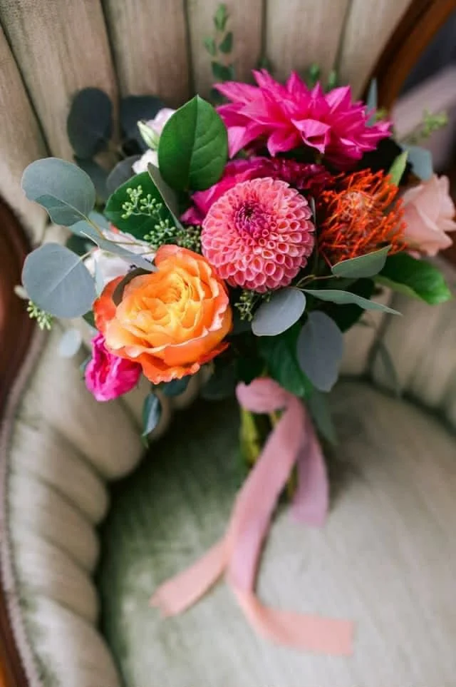 Colorful bouquet of pink, orange, and purple flowers with green leaves and a pink ribbon, placed on a vintage-style chair with a wooden background.