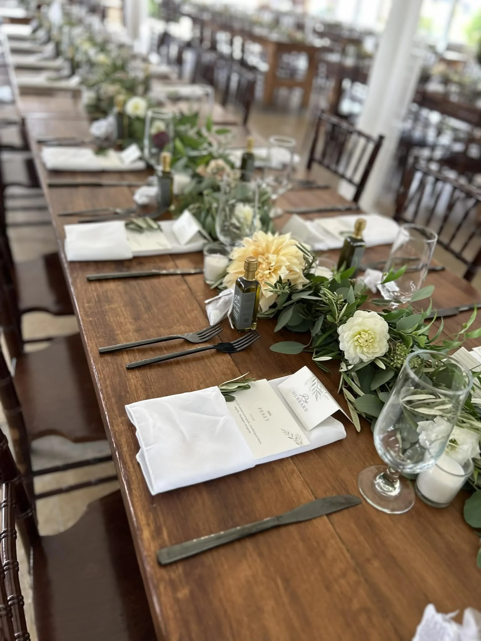 Long wooden banquet table set for a formal event with white cloth napkins, black utensils, small bottles of olive oil, greenery and cream-colored floral centerpieces, glassware, and printed menus.