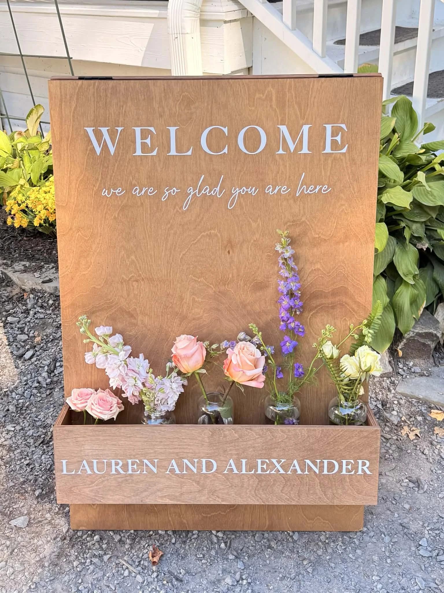 Wooden welcome sign with flowers in small glass vases in front, reading 'Welcome, we are so glad you are here, Lauren and Alexander' surrounded by greenery and flowers lovingly created for the bride and groom by Lot 32 Flower Farm.