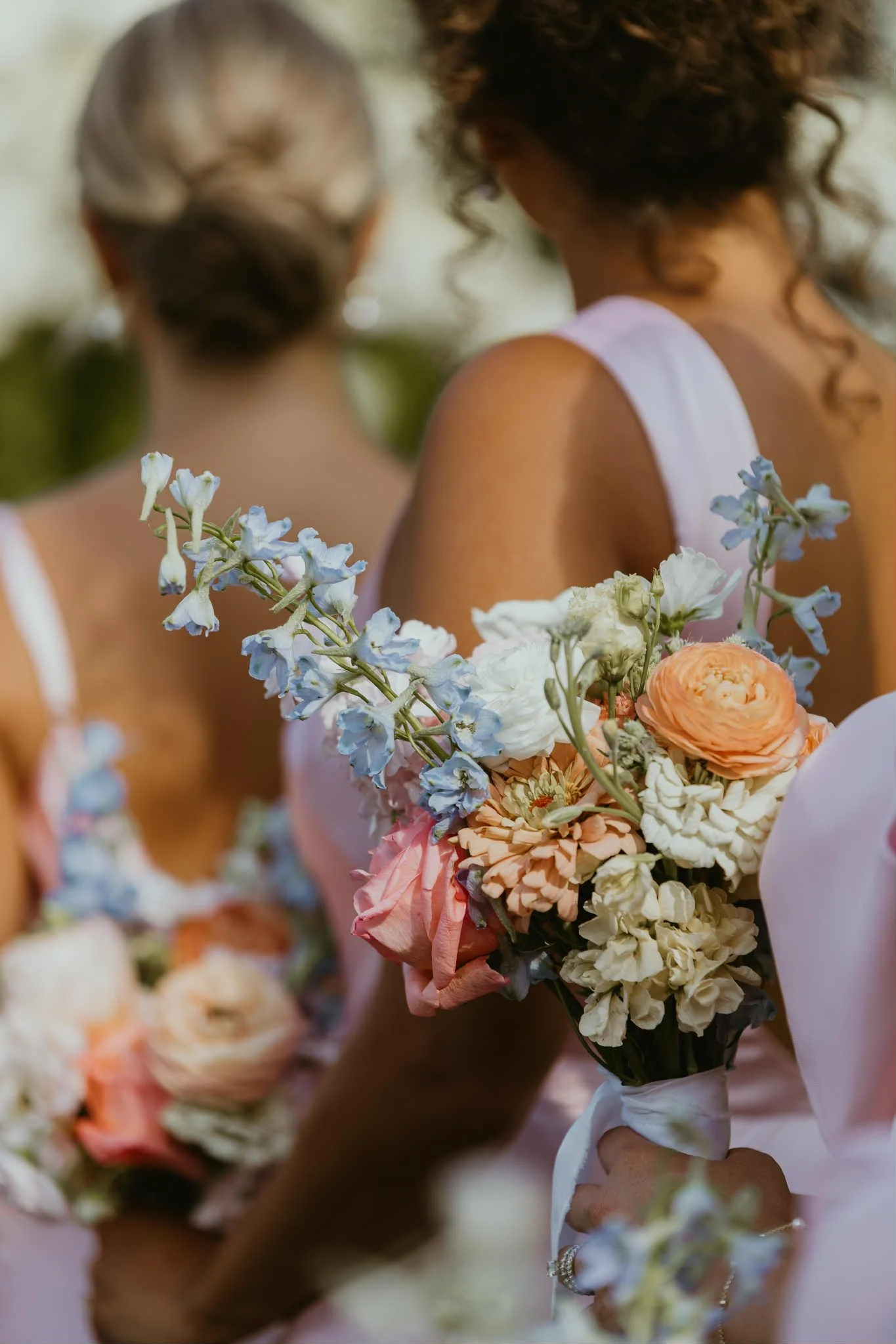 Close-up of a bouquet of mixed flowers held by a woman at a wedding, with other women and similar bouquets visible in the blurred background.