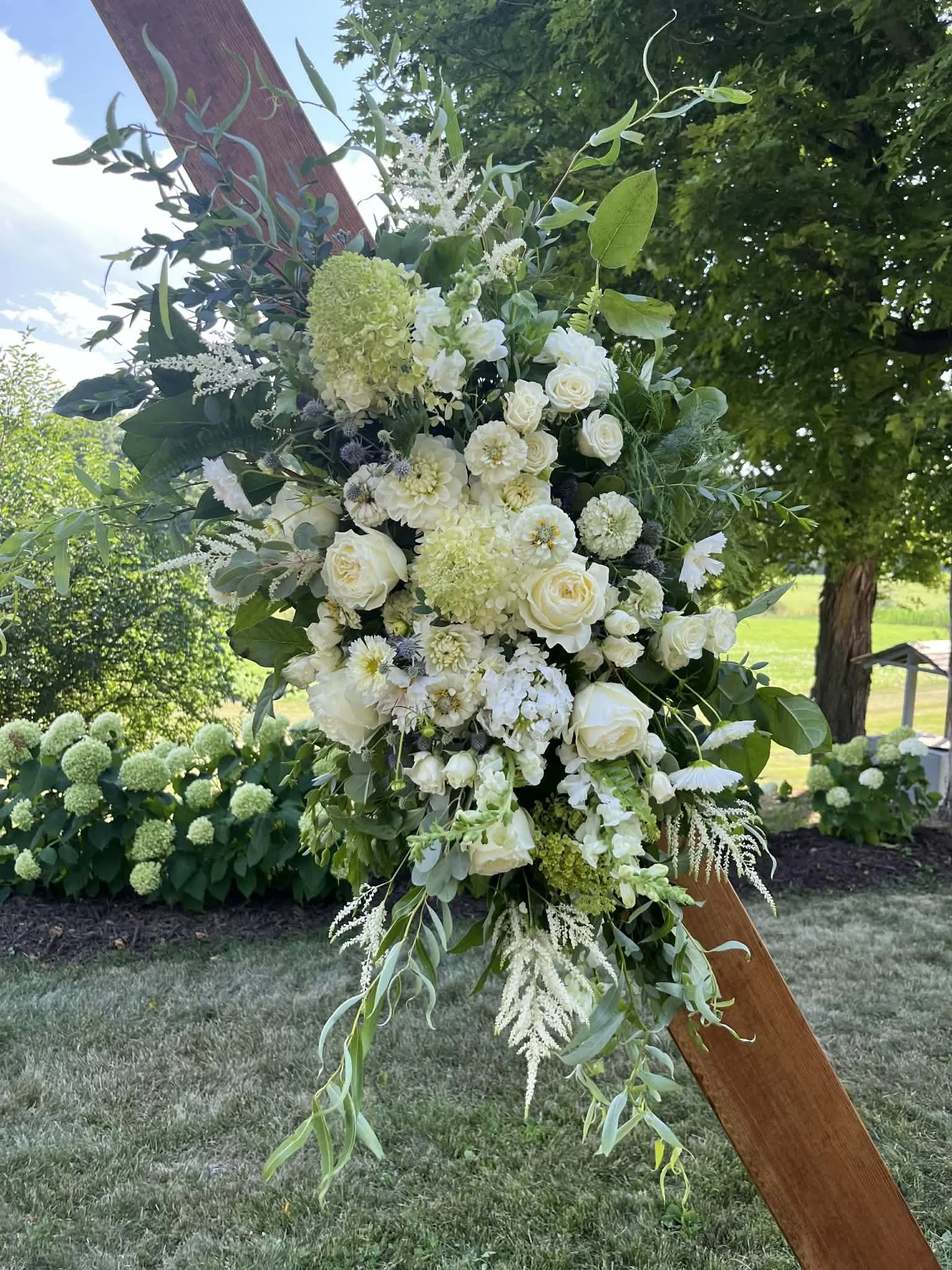 White floral arrangement with roses, hydrangeas, and greenery attached to a wooden structure outdoors.