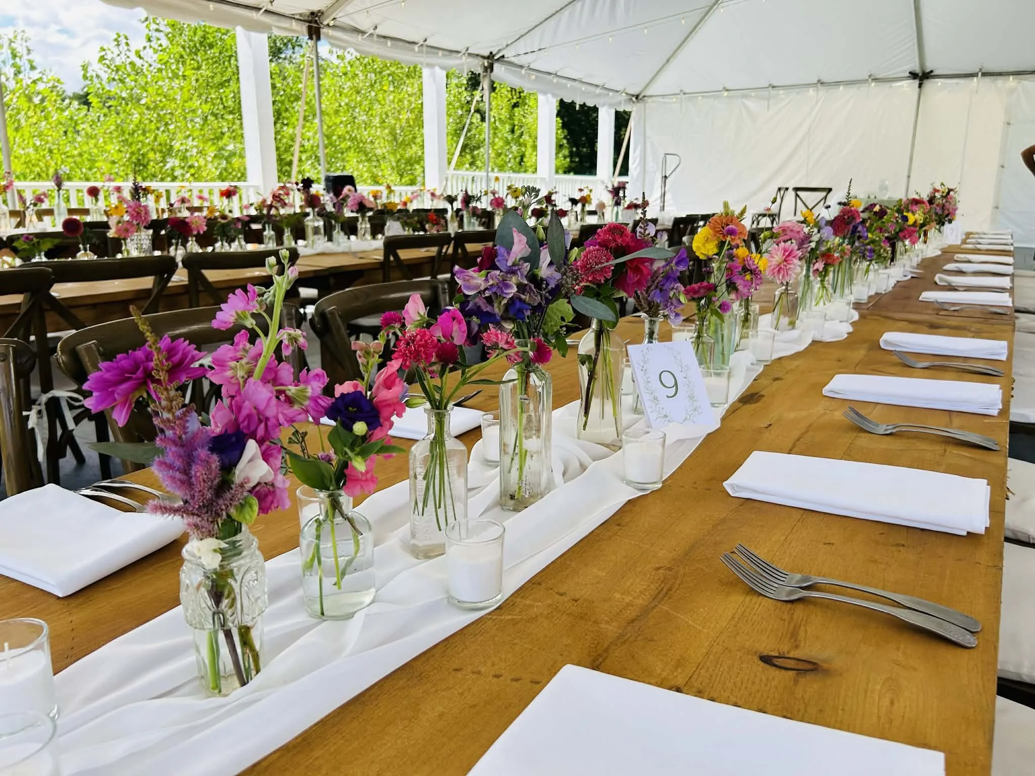 Long wooden dining table decorated with colorful flower arrangements in glass vases, white cloth napkins, forks, and candles under a white tent, with green foliage visible outside.