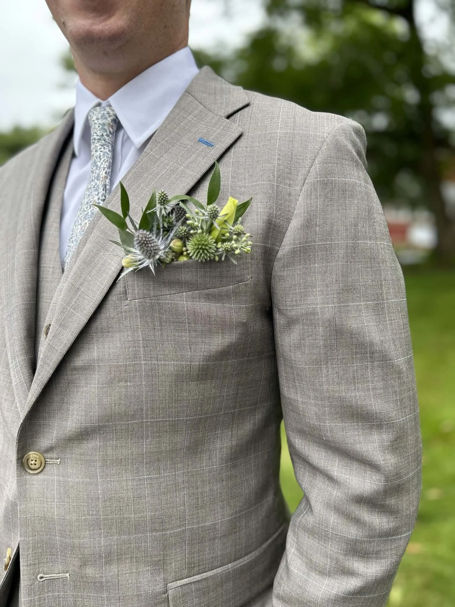 Man wearing a light grey checkered suit with a white shirt and floral tie, with a boutonniere of green and yellow flowers and leaves on his lapel, outdoors with trees in the background.