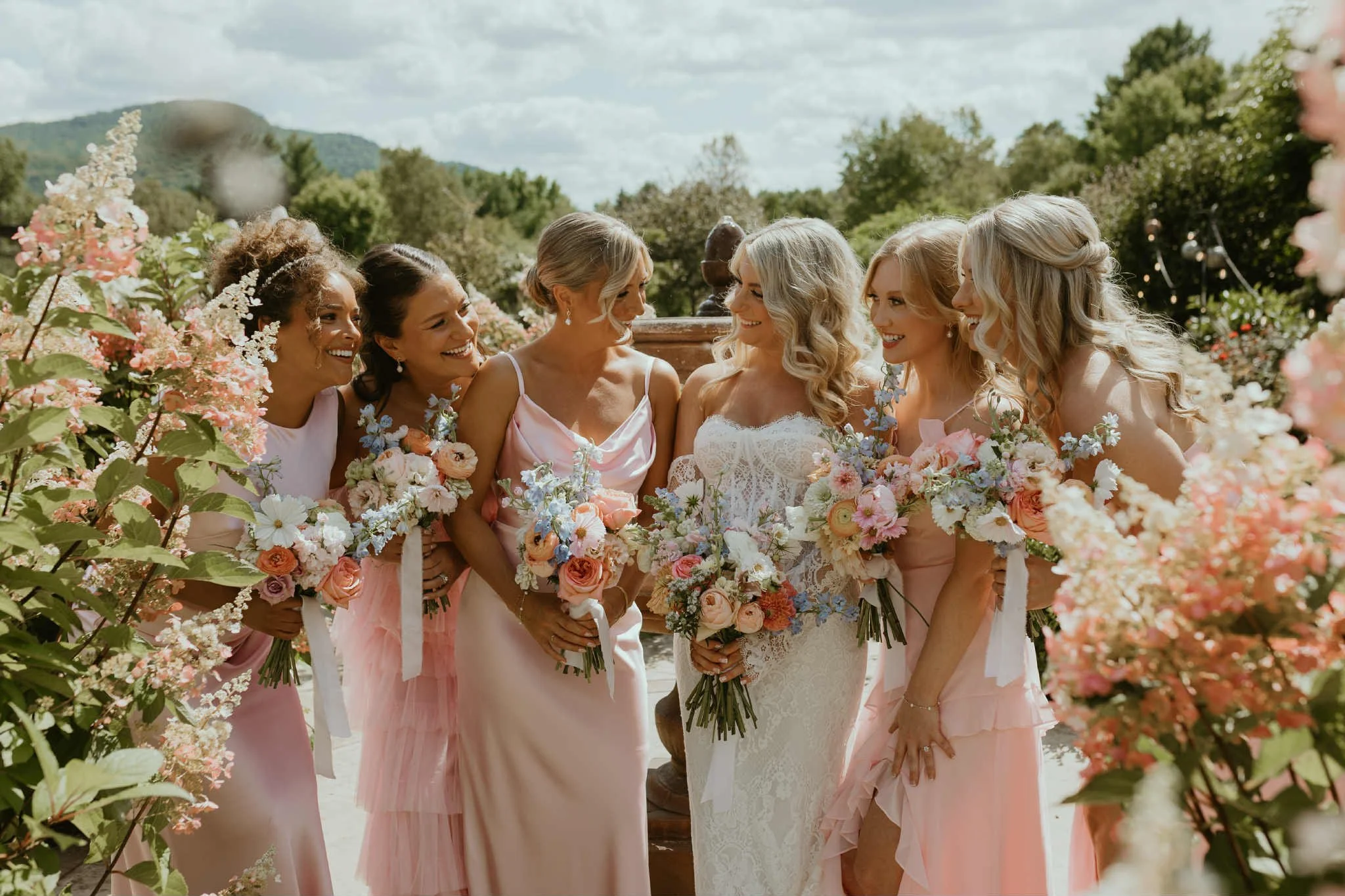 Group of women in pastel dresses, holding bouquets, smiling and interacting outdoors during a wedding.