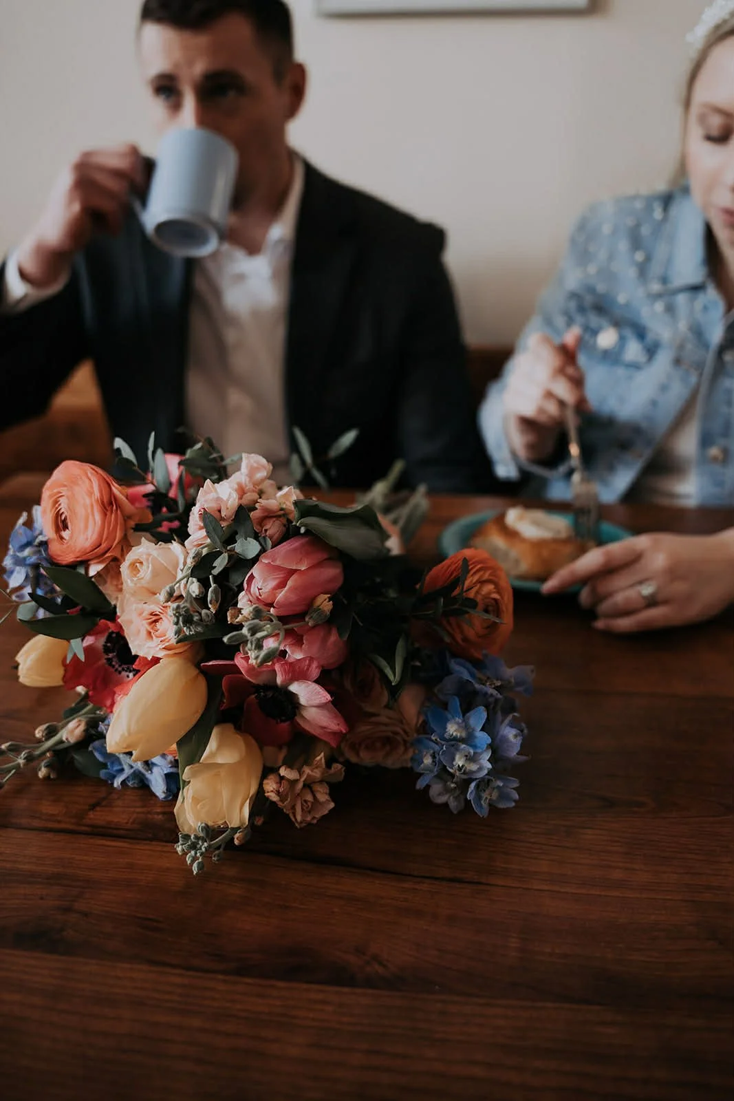 A bouquet of colorful flowers on a wooden table with a man drinking from a gray mug and a woman eating cake in the background.