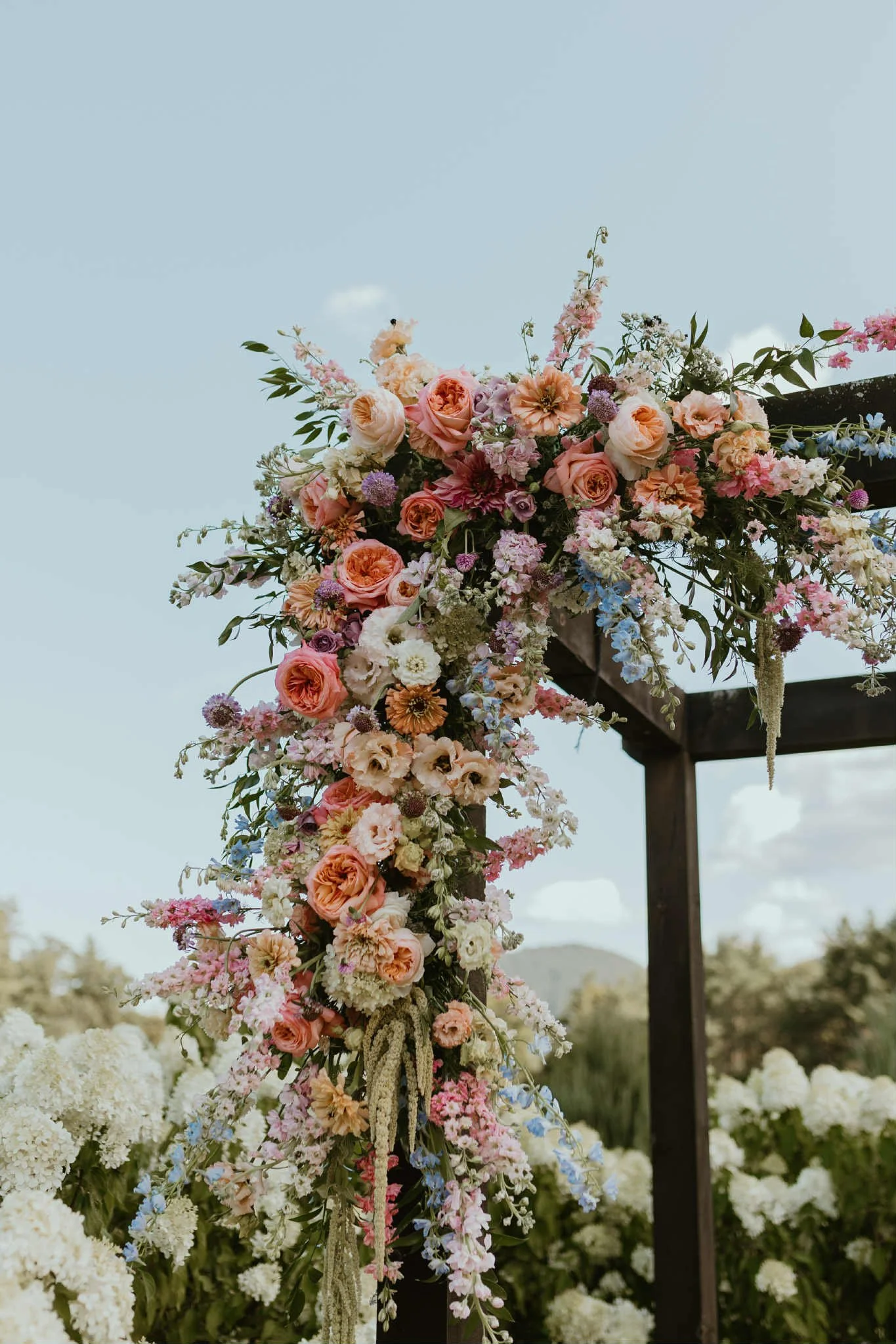 A floral arbor created by Lot 32 Flower Farm with pink, white, purple, and peach flowers hanging from a black wooden frame outdoors under a partly cloudy sky.