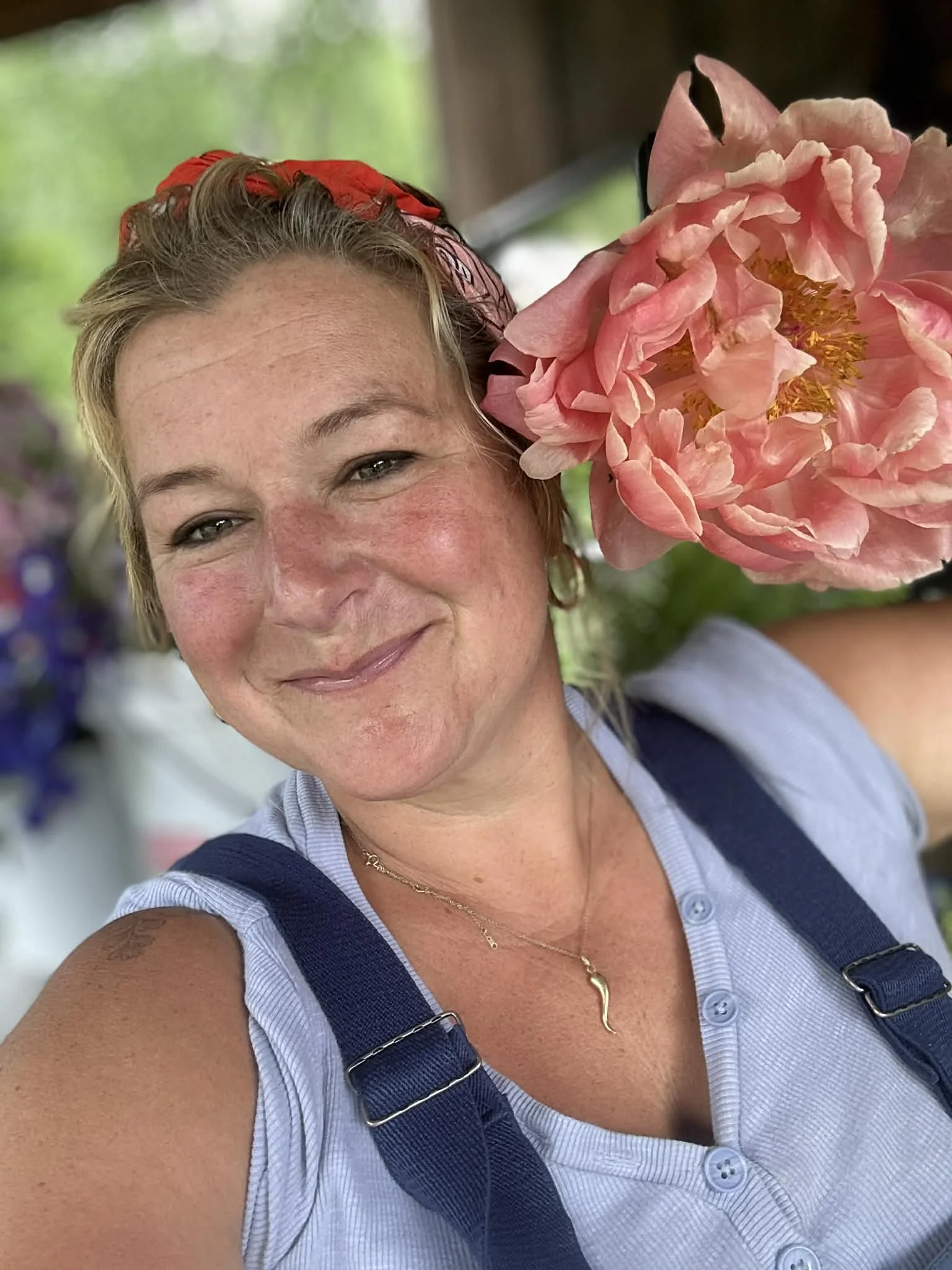 A woman taking a selfie outdoors, holding a large pink flower close to her face. She has short blonde hair, is smiling, and is wearing a blue sleeveless shirt, a gold necklace, and a red headband.