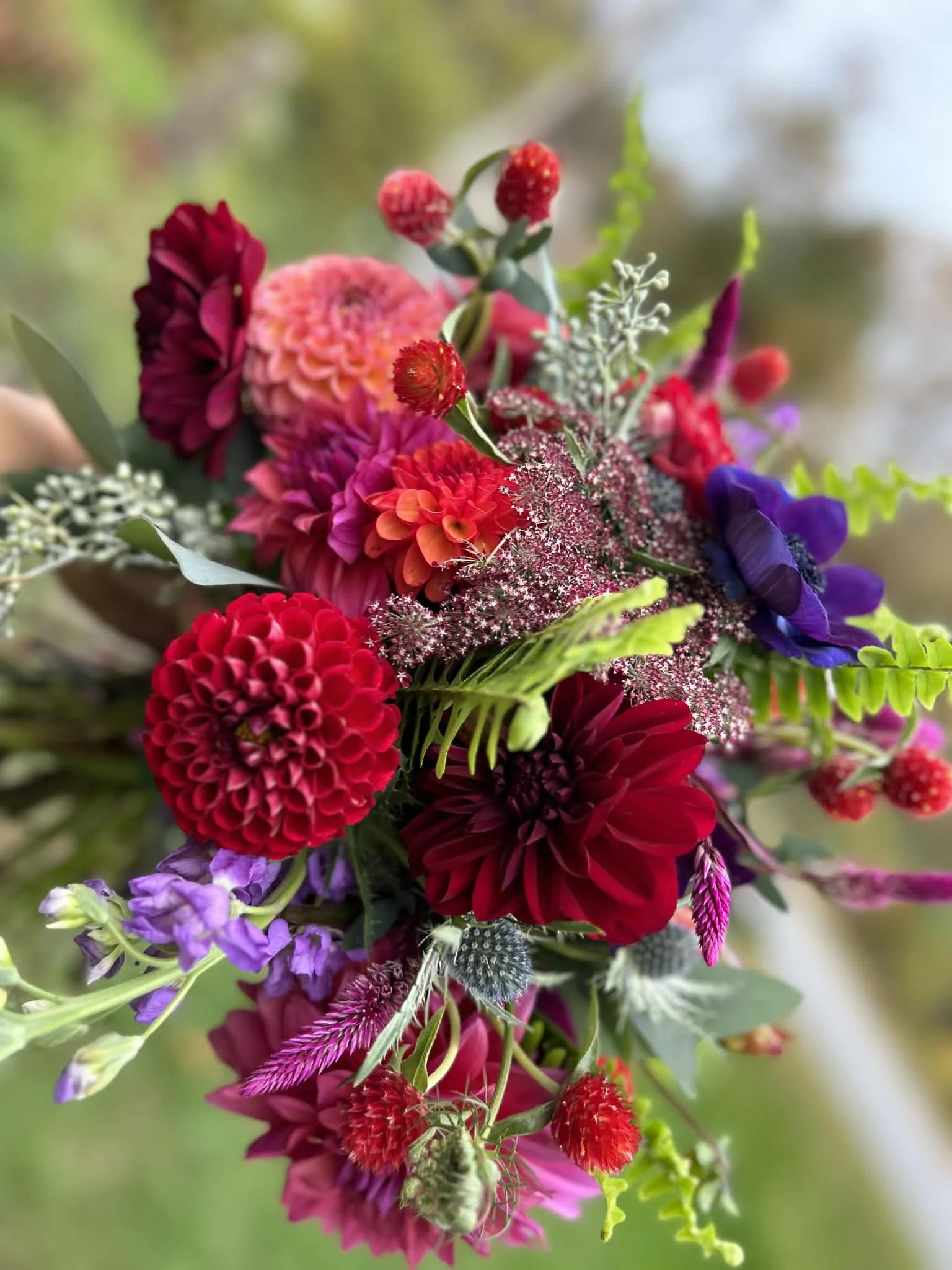 A colorful bouquet of various flowers including dahlias, and other vibrant blossoms in shades of red, pink, purple, and orange on a blurred natural background.
