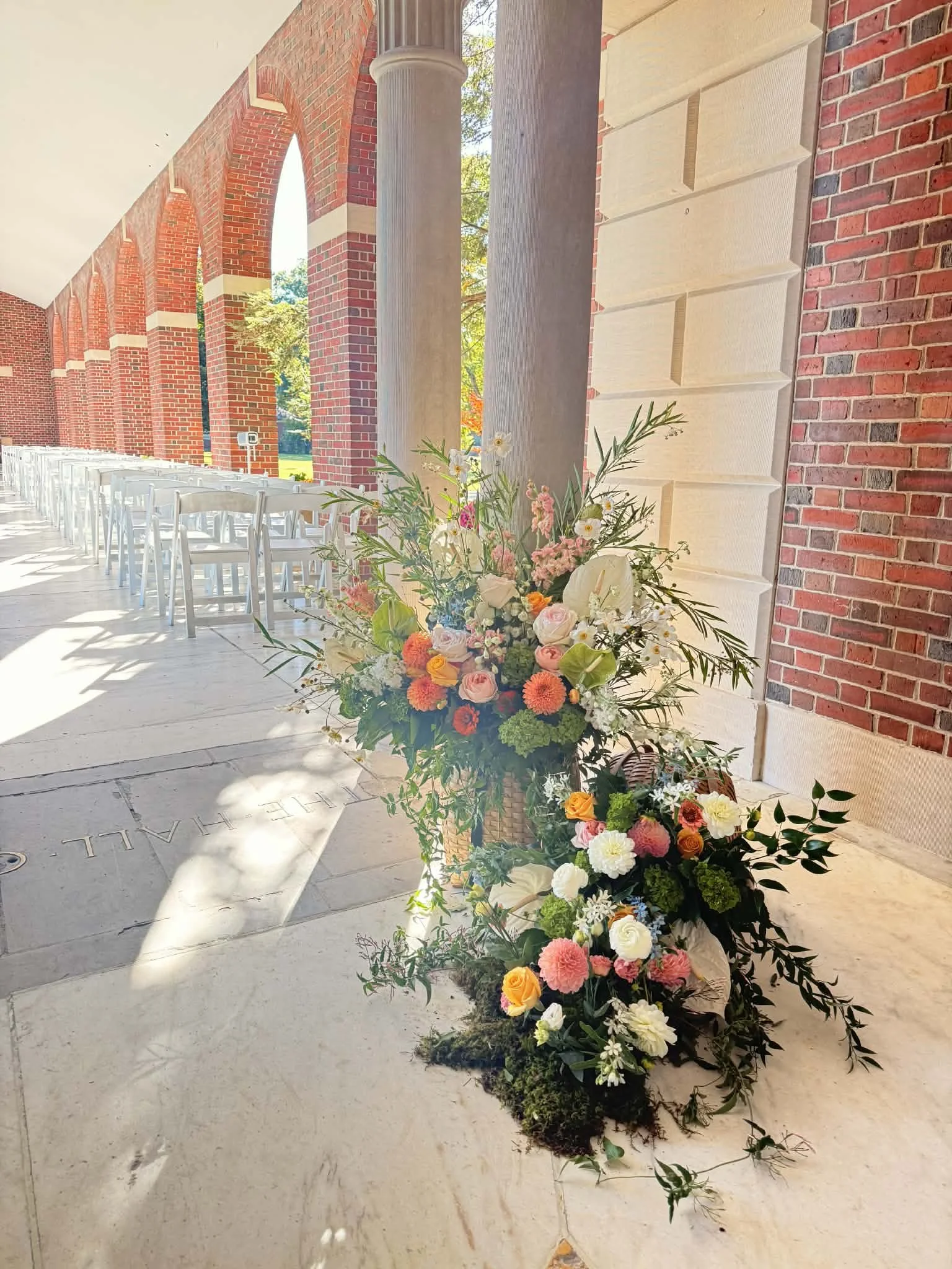 A floral arrangement with pink, white, orange, and green flowers and greenery placed on the ground near a brick wall with tall columns and an outdoor archway at a venue decorated for an event.