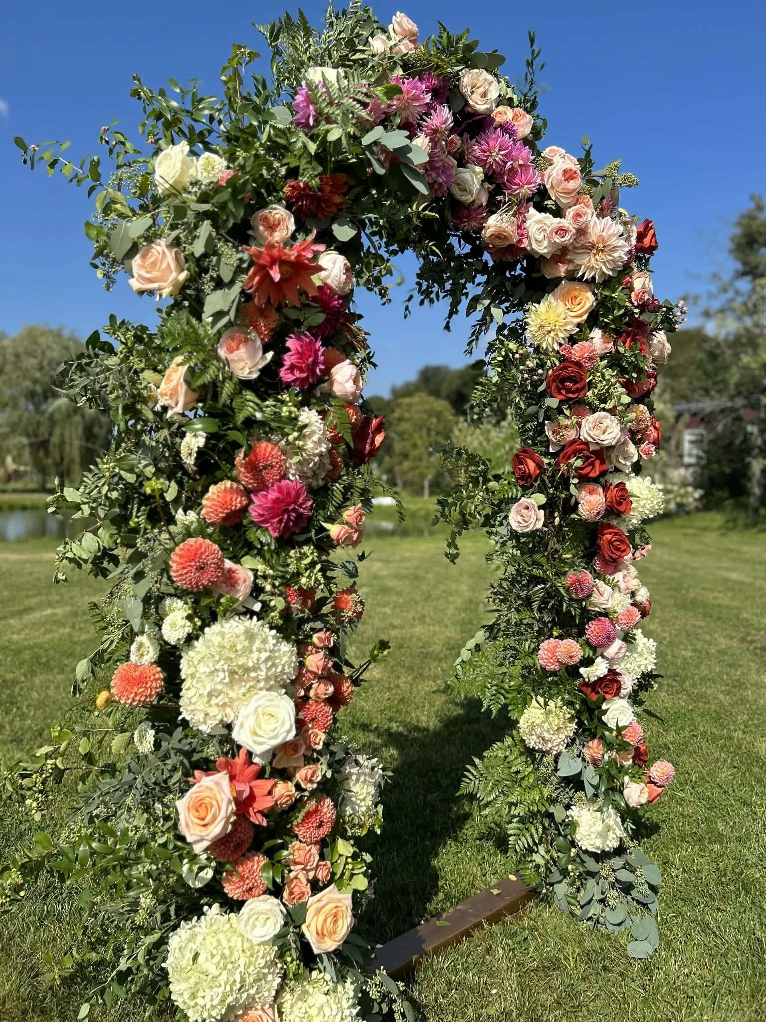 A large floral arch made of various pink, white, peach, and red flowers with green foliage created by Lot 32 Flower Farm outdoors on a sunny day with a field of green grass in the background.