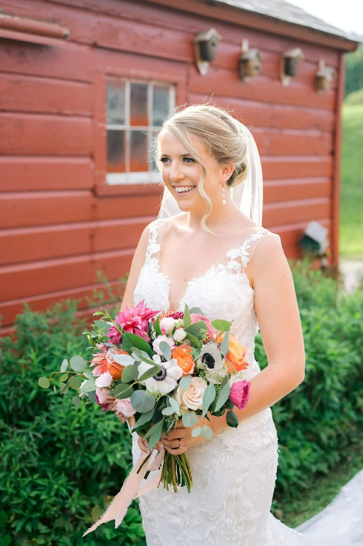 A bride in a white lace wedding gown holding a colorful bouquet of flowers outdoors near a red wooden building.