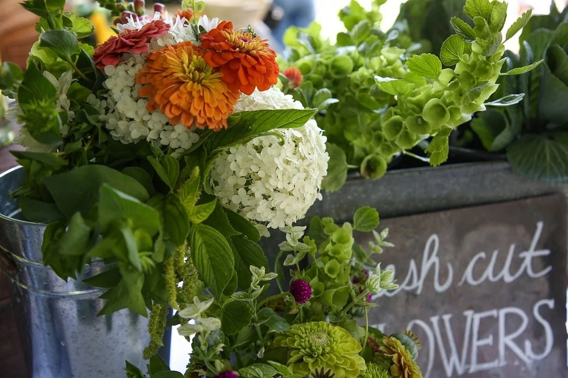 Arrangement of colorful flowers, including white hydrangeas, orange and pink zinnias, and green foliage in a metallic container with a chalkboard sign reading 'fresh cut flowers' in the background.