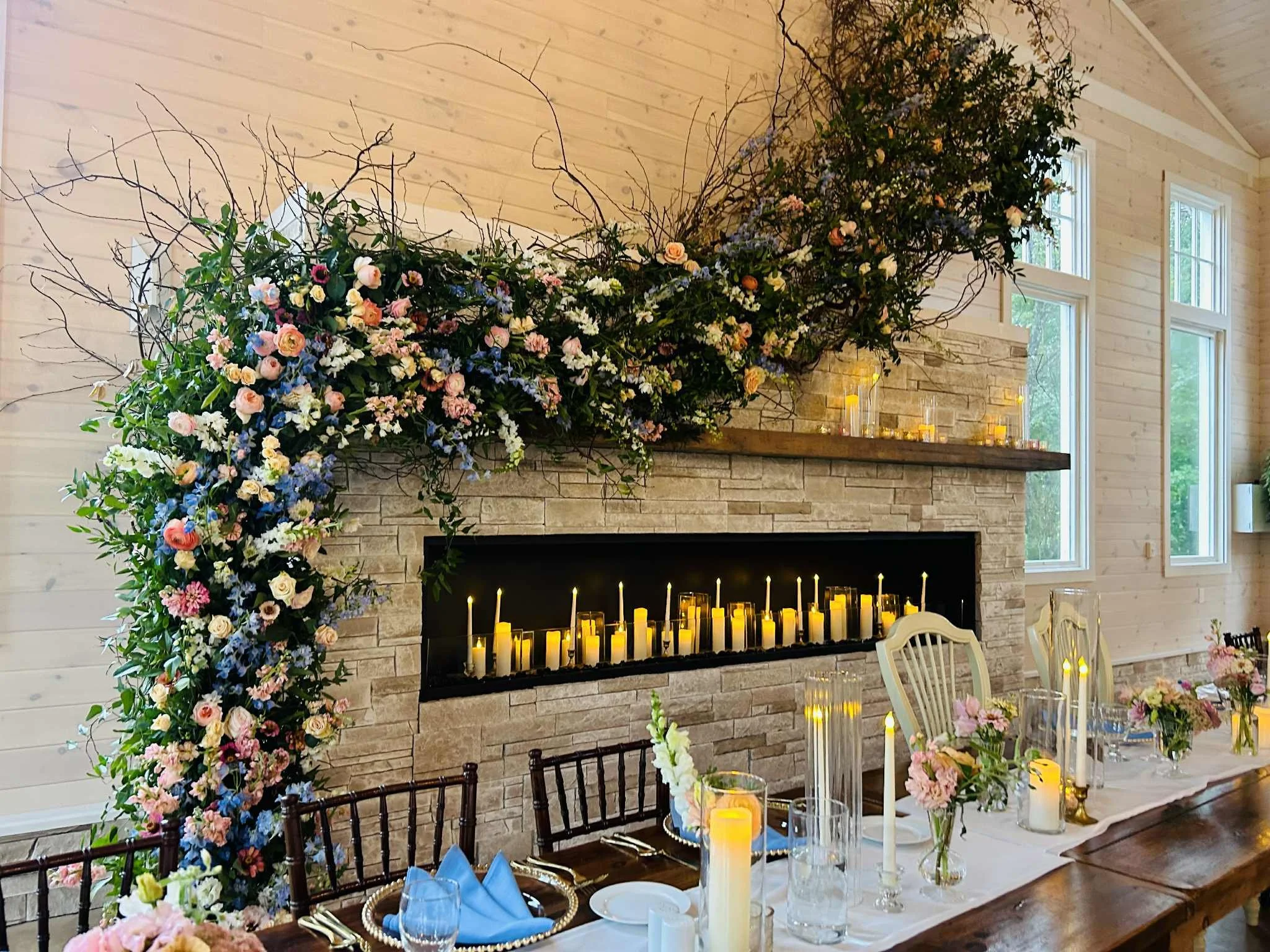 A decorated fireplace with a lush floral arrangement and candles, set in a dining area with windows and a long table.