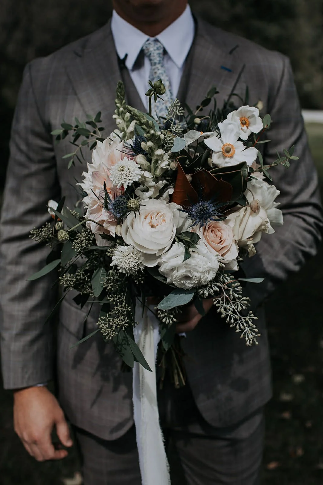 Person in a gray plaid suit holding a bouquet of white and light pink flowers, including roses, peonies, and greenery, with a white ribbon.