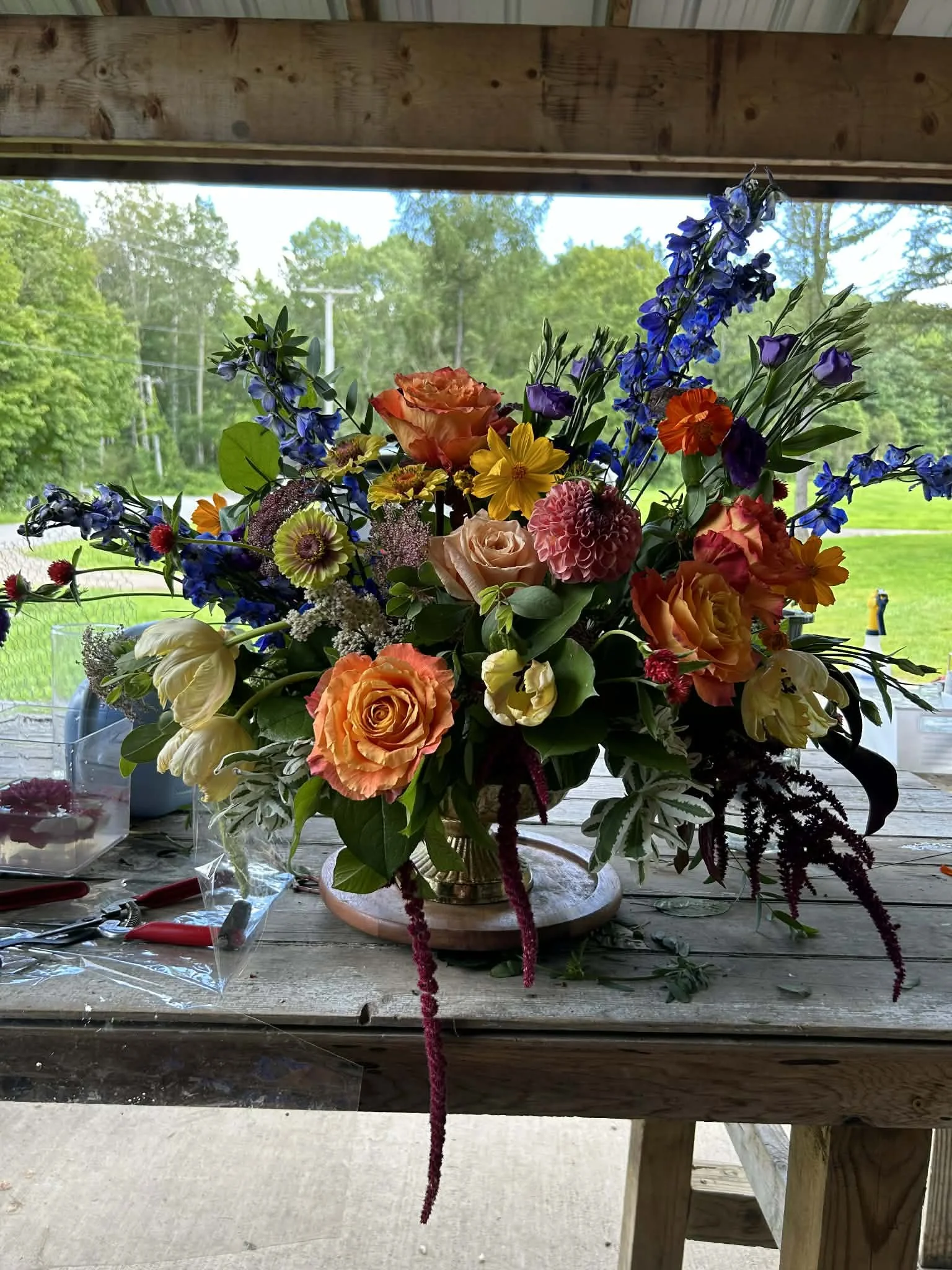 A colorful bouquet of flowers on a wooden table, with various flowers including roses, delphiniums, and dahlias in a vase, set outdoors with green trees and a grassy field in the background.