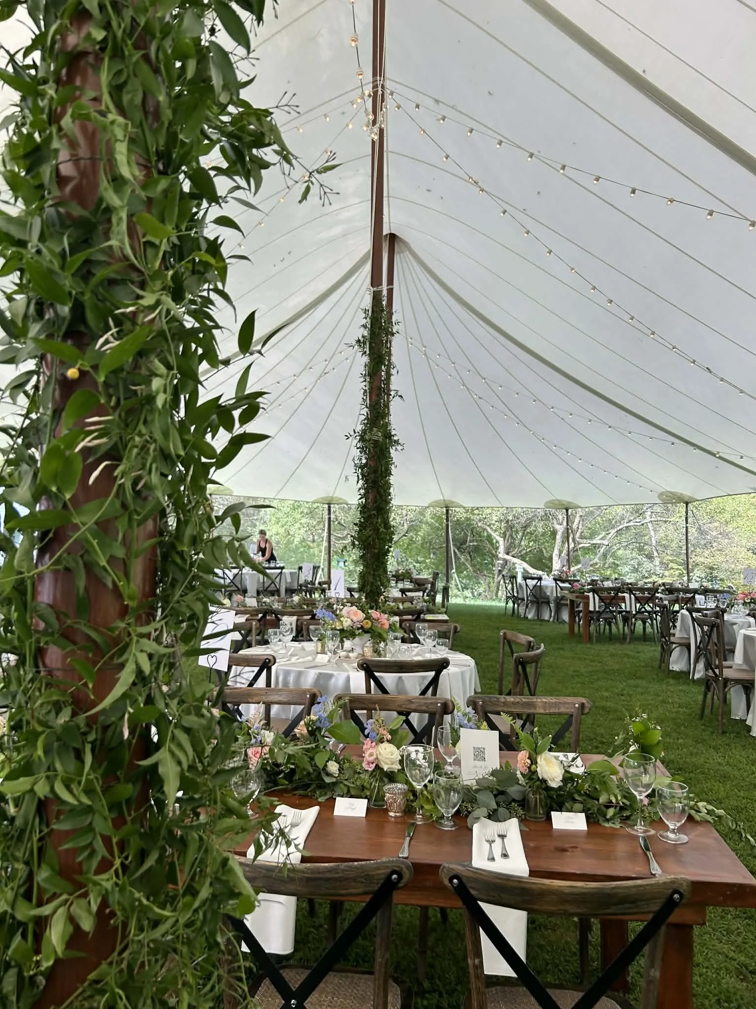 Inside a decorated outdoor event tent with tables set for a celebration, adorned with floral centerpieces and fairy lights, surrounded by greenery and trees in the background.