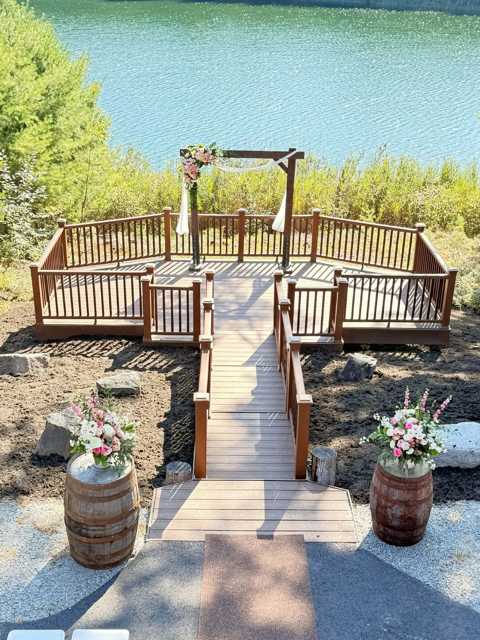 A wooden outdoor wedding altar decorated with pink and white flowers, set on a platform overlooking a body of water, with flower arrangements in barrel planters on either side.