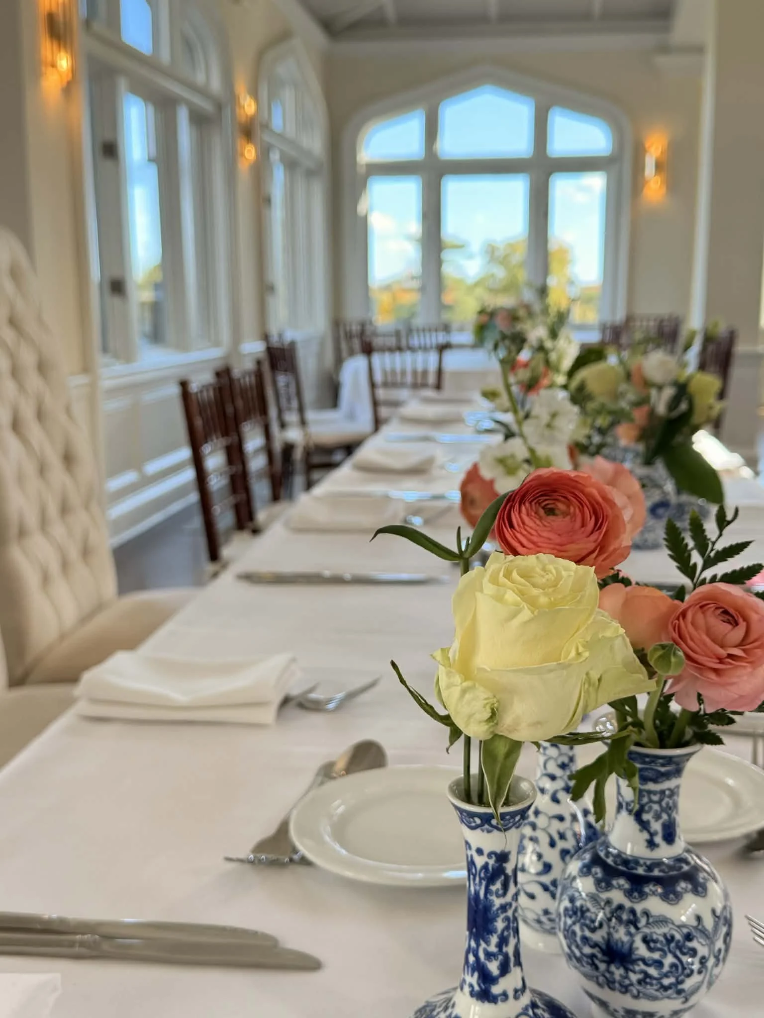 Close-up of a dining table with floral centerpieces in blue-and-white vases, set with white plates, silverware, and folded napkins, in a bright room with large windows and natural light.