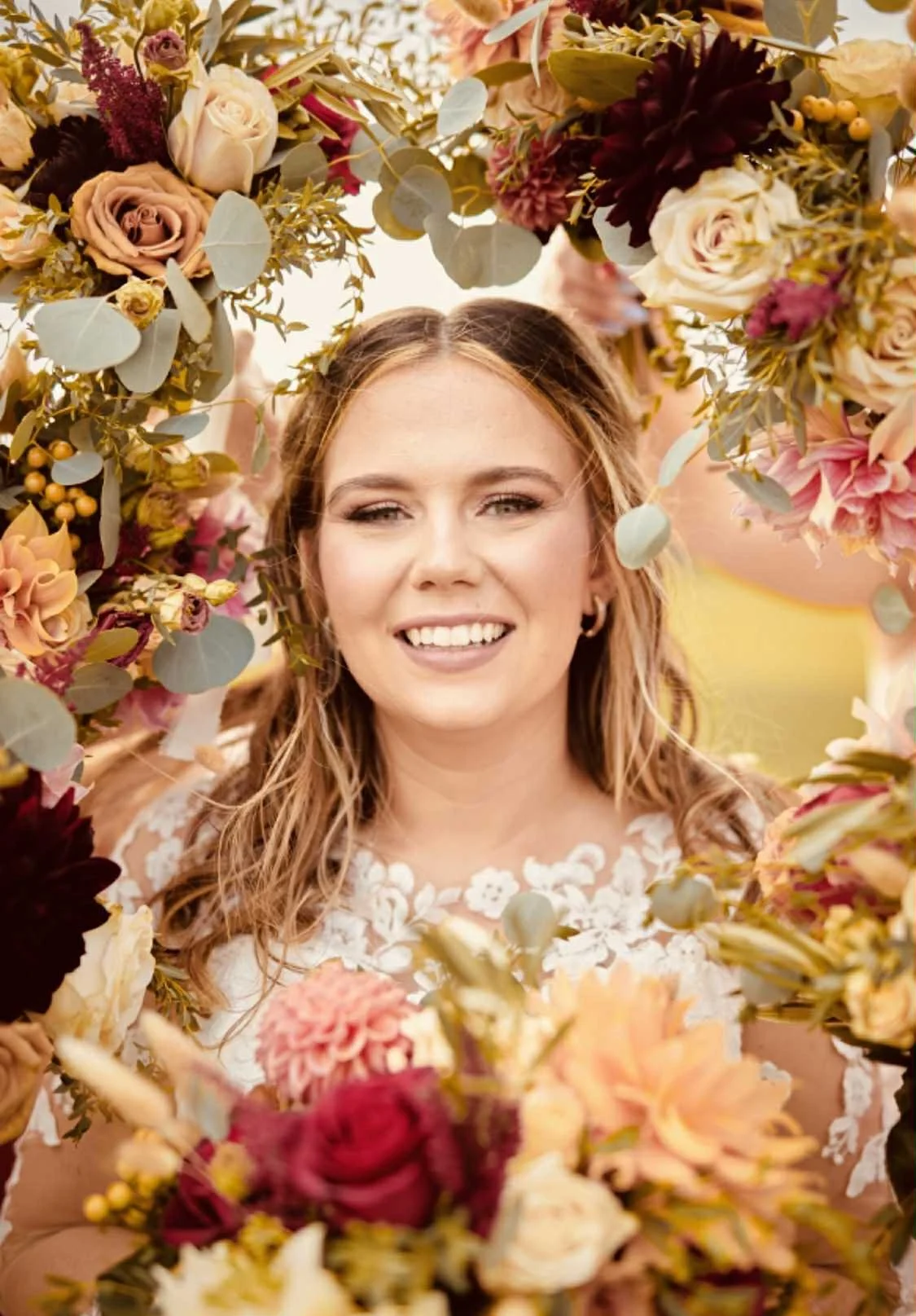 A smiling woman with wavy blonde hair, surrounded by a floral wedding arch with pink, white, and burgundy flowers and green foliage.