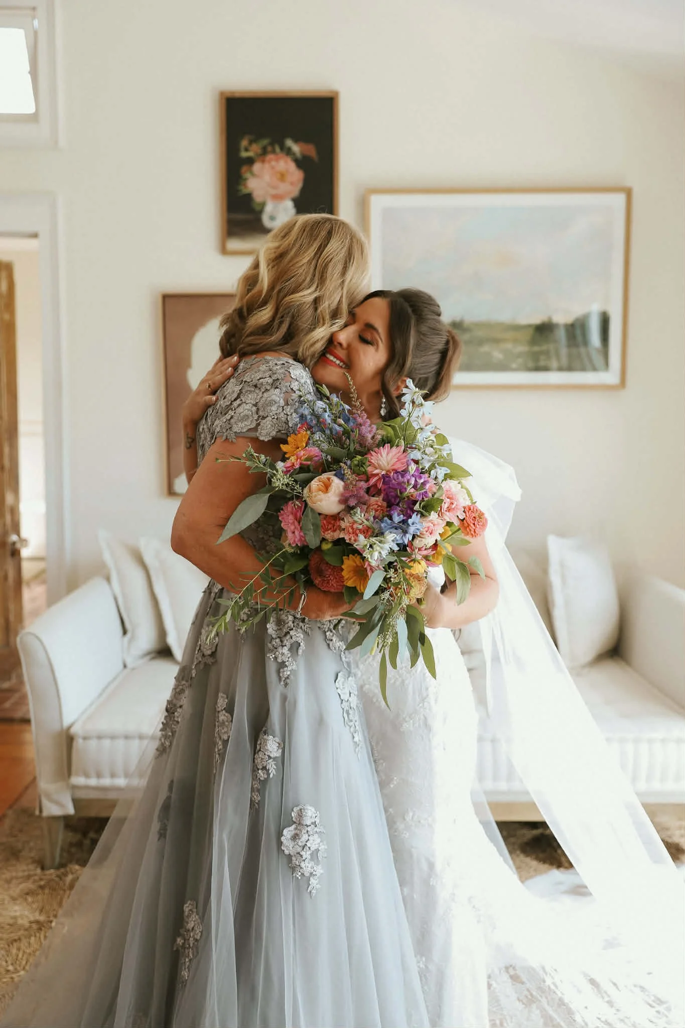 Two women hugging, one in a wedding dress holding a colorful bouquet, in a room with white walls, artwork, and a sofa.