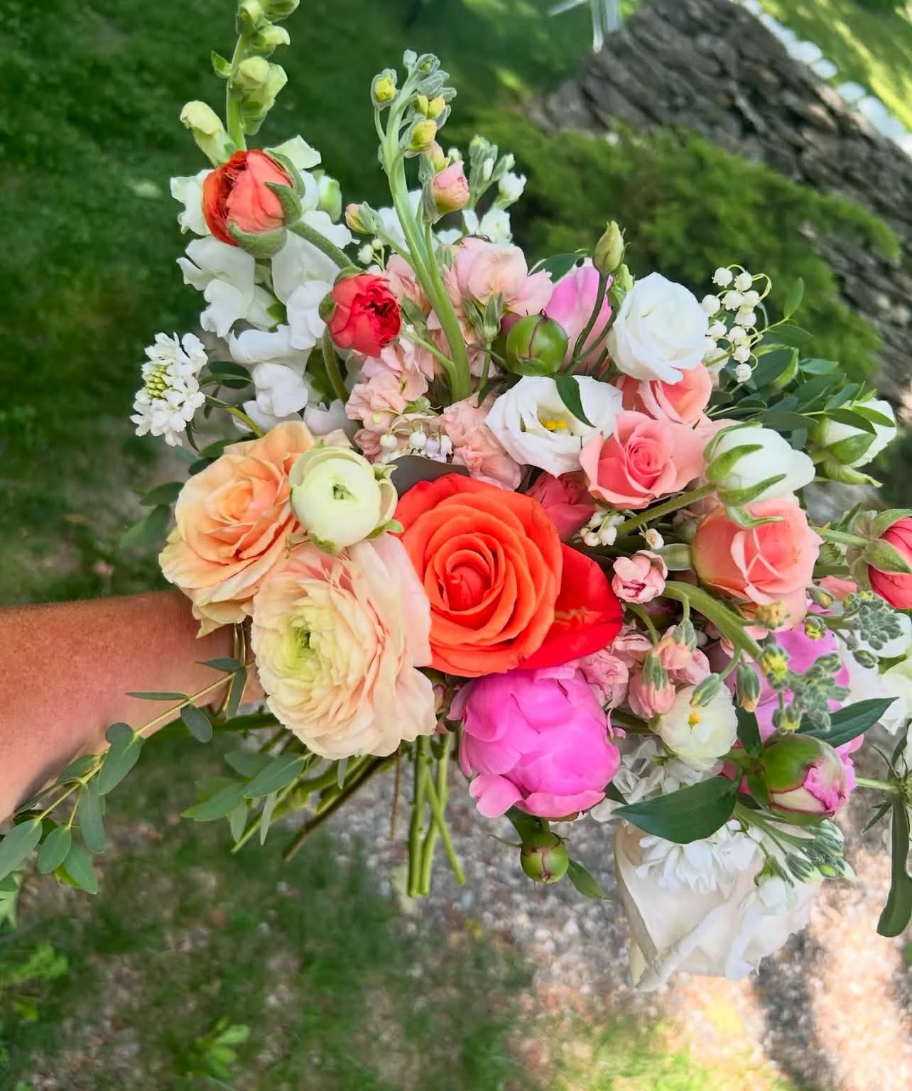 A colorful bouquet of various flowers including roses, lisianthus, and other mixed blooms, held outdoors with a tree and grass in the background.