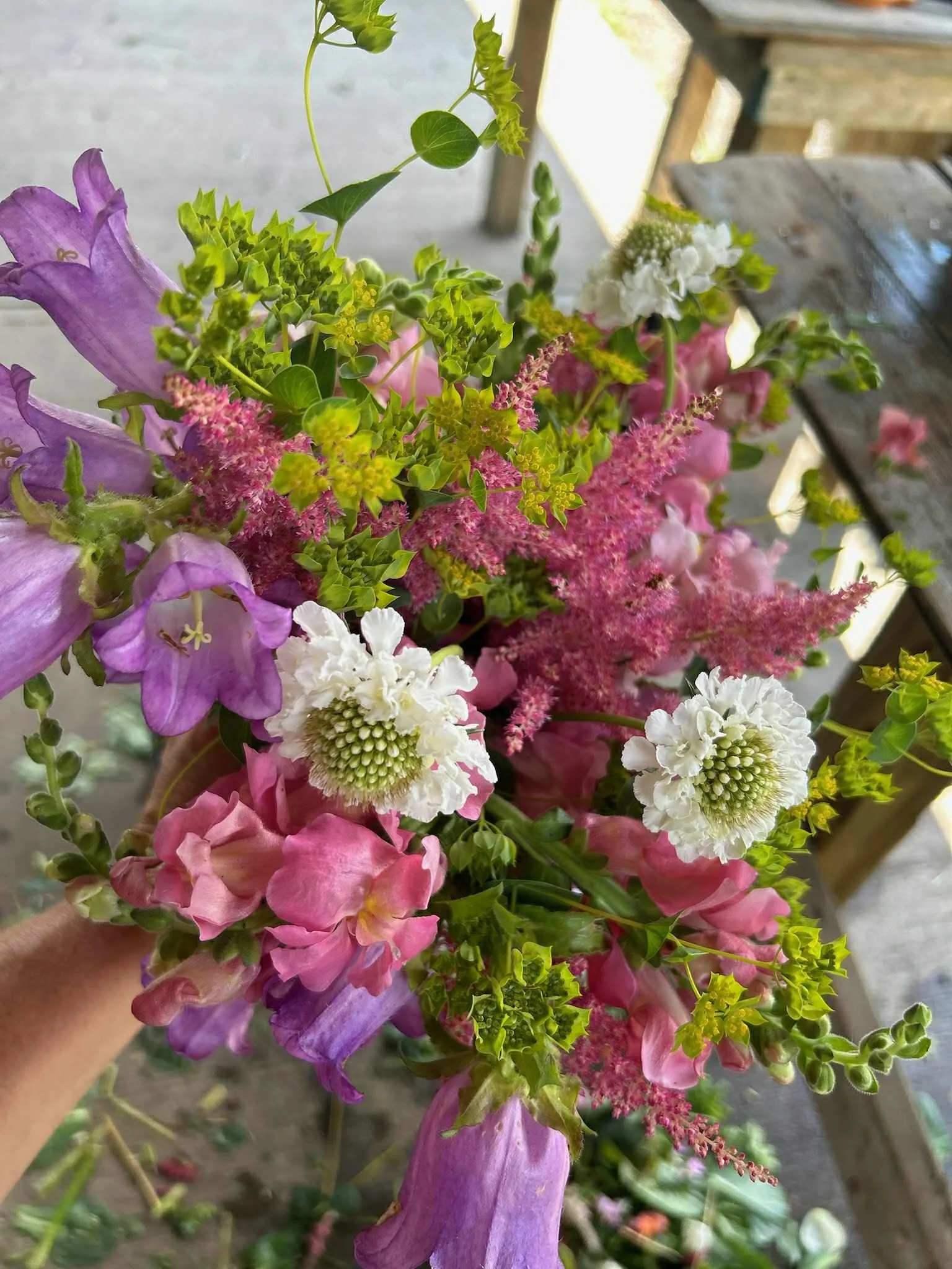 A colorful bouquet of various flowers including purple, pink, and white blooms, with green leaves, held by a hand.