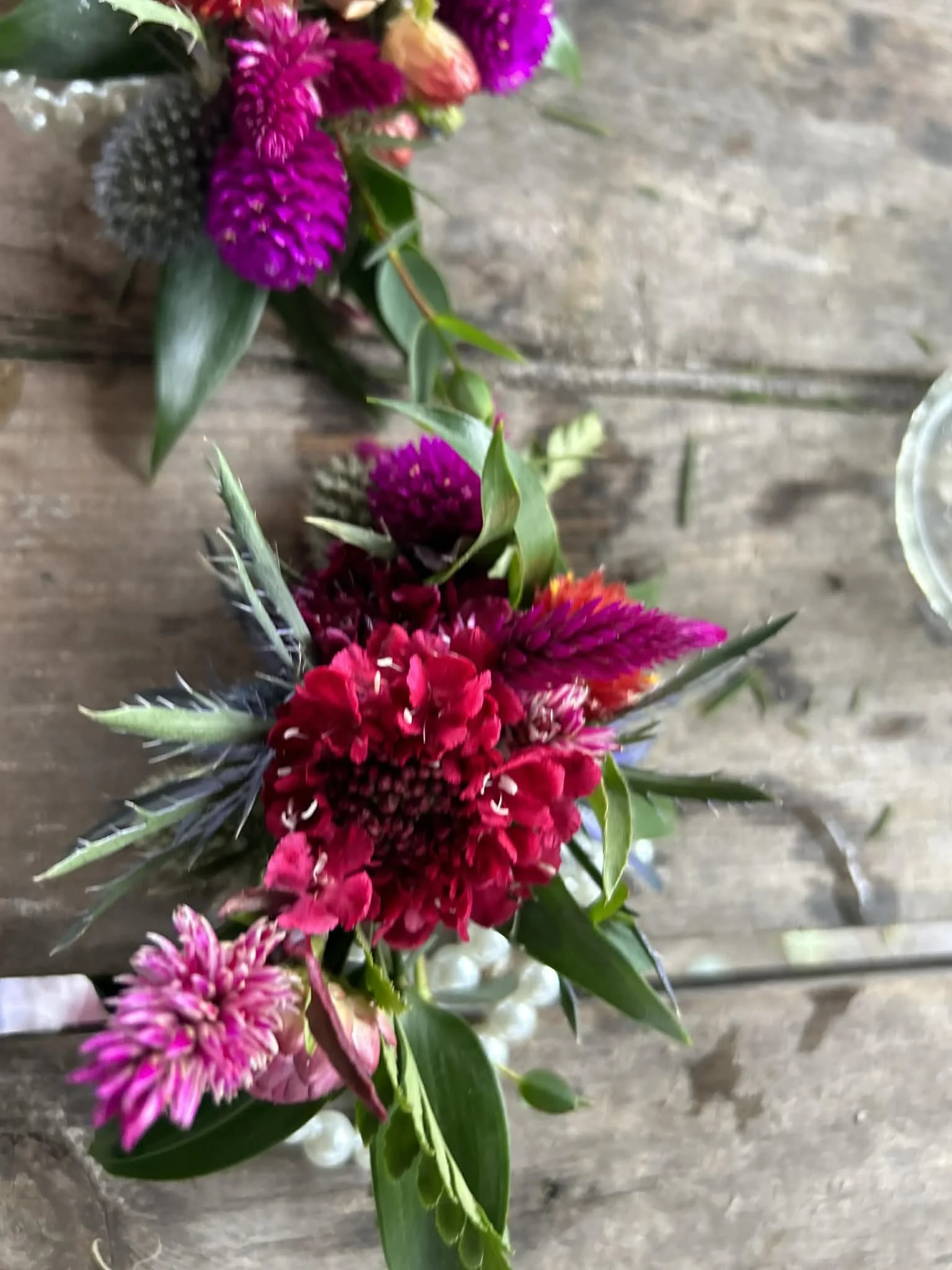 Colorful braceket corsage for the mothers of the bride and groom designed by Lot 32 Flower Farm with pink, purple, and red flowers, surrounded by green leaves, placed on a rustic wooden surface.